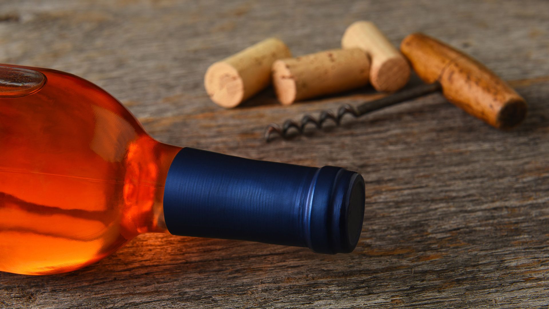 A close-up shot of a bottle of blush wine with a dark blue label, lying on its side on a rustic wooden surface. In the background, out of focus, are several wine corks and a corkscrew.