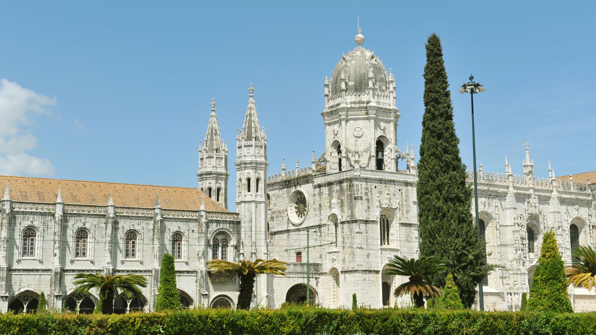 A grand, ornate white stone monastery with towering spires and a large dome, set against a clear blue sky with green foliage and palm trees in the foreground.