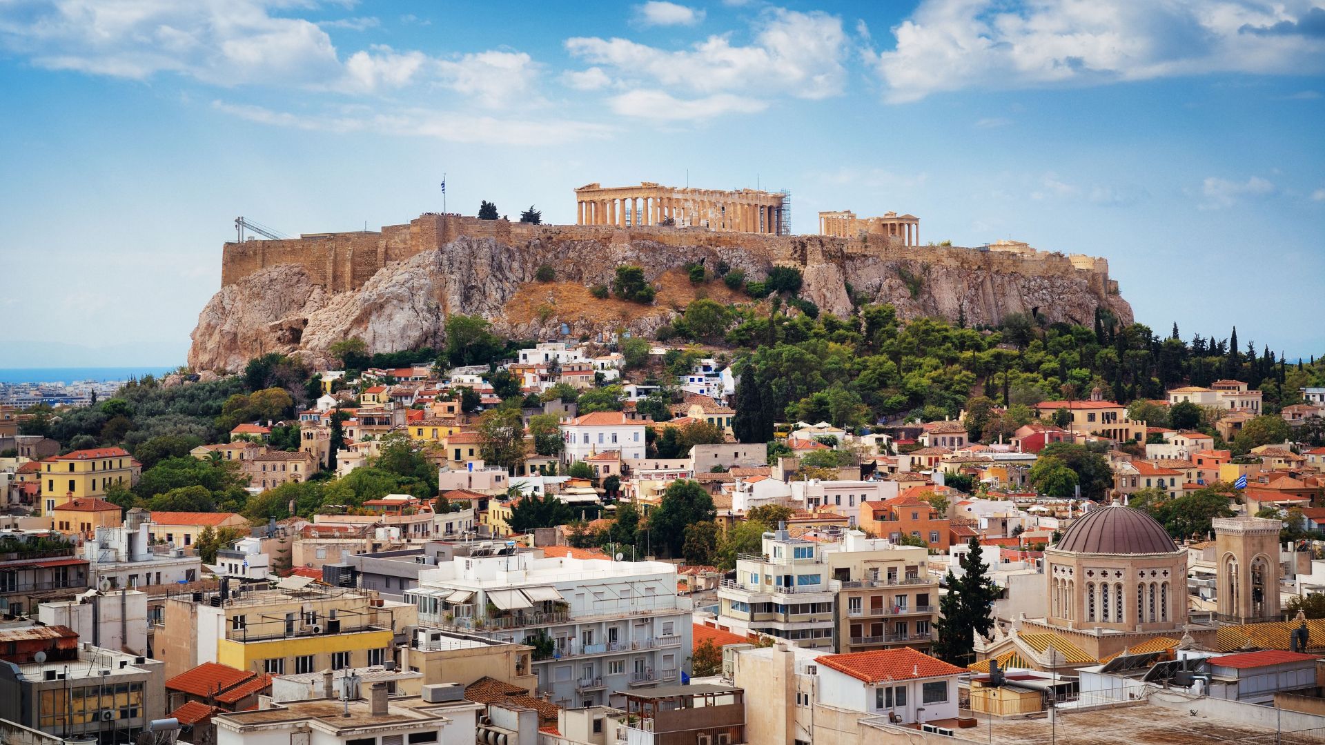 A panoramic view of the ancient Parthenon temple atop the rocky Acropolis hill, overlooking the densely built-up city of Athens under a clear sky with scattered clouds.
