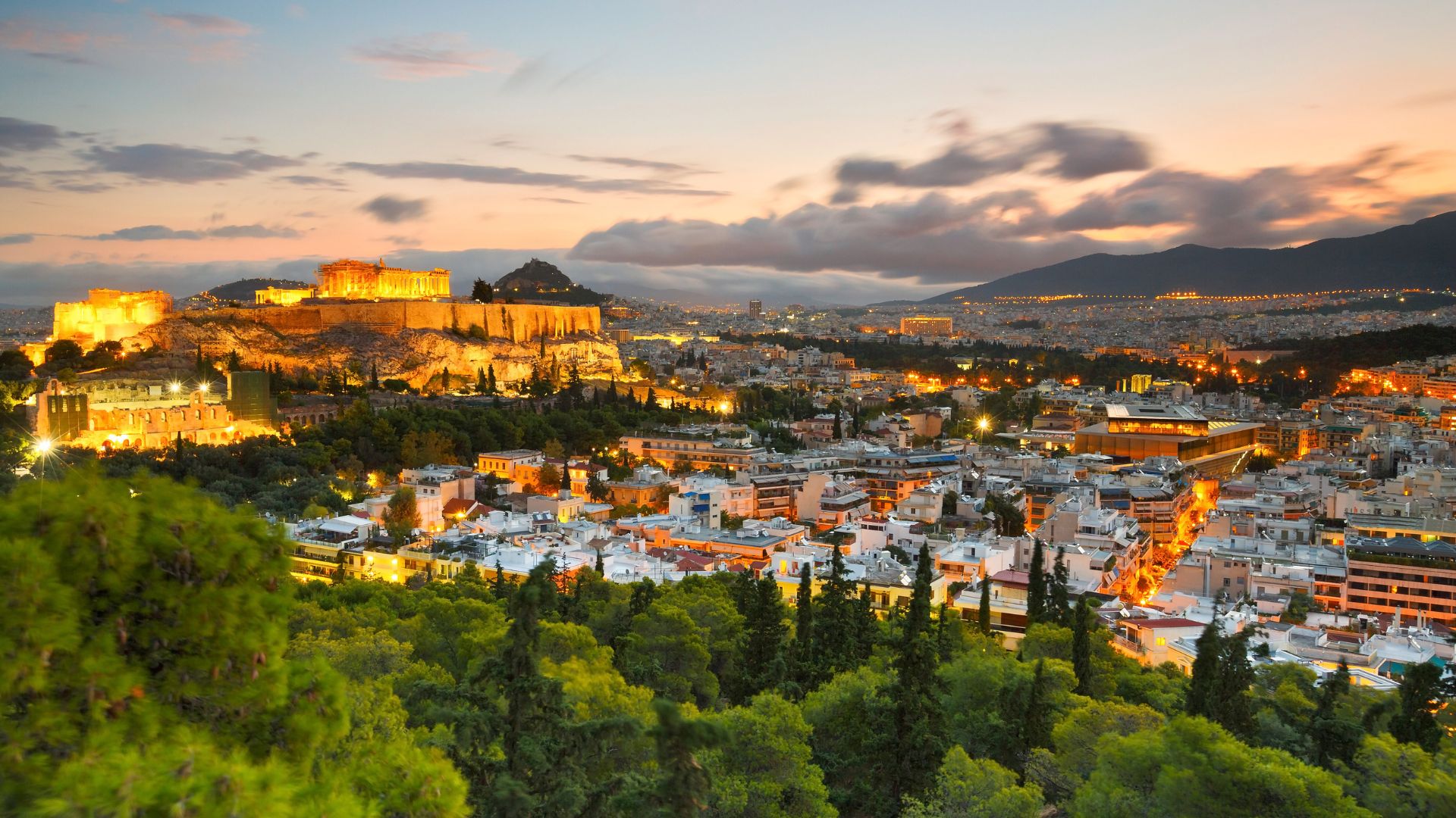 A panoramic view of the illuminated Acropolis of Athens at dusk, with the ancient citadel atop a hill overlooking the sprawling city lights and surrounding mountains under a dramatic sky.