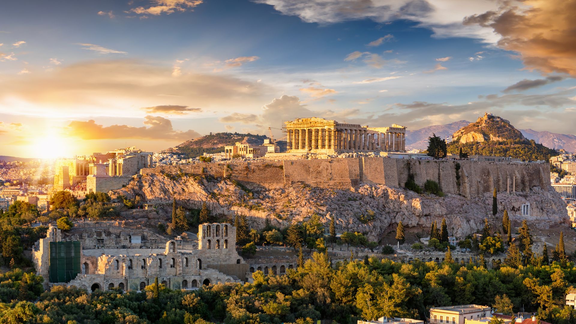 A wide-angle shot at sunset of the ancient Acropolis in Athens, Greece, showcasing the majestic Parthenon temple atop the hill, with the Odeon of Herodes Atticus visible in the foreground, surrounded by lush greenery and the city extending into the distance under a vibrant sky. 