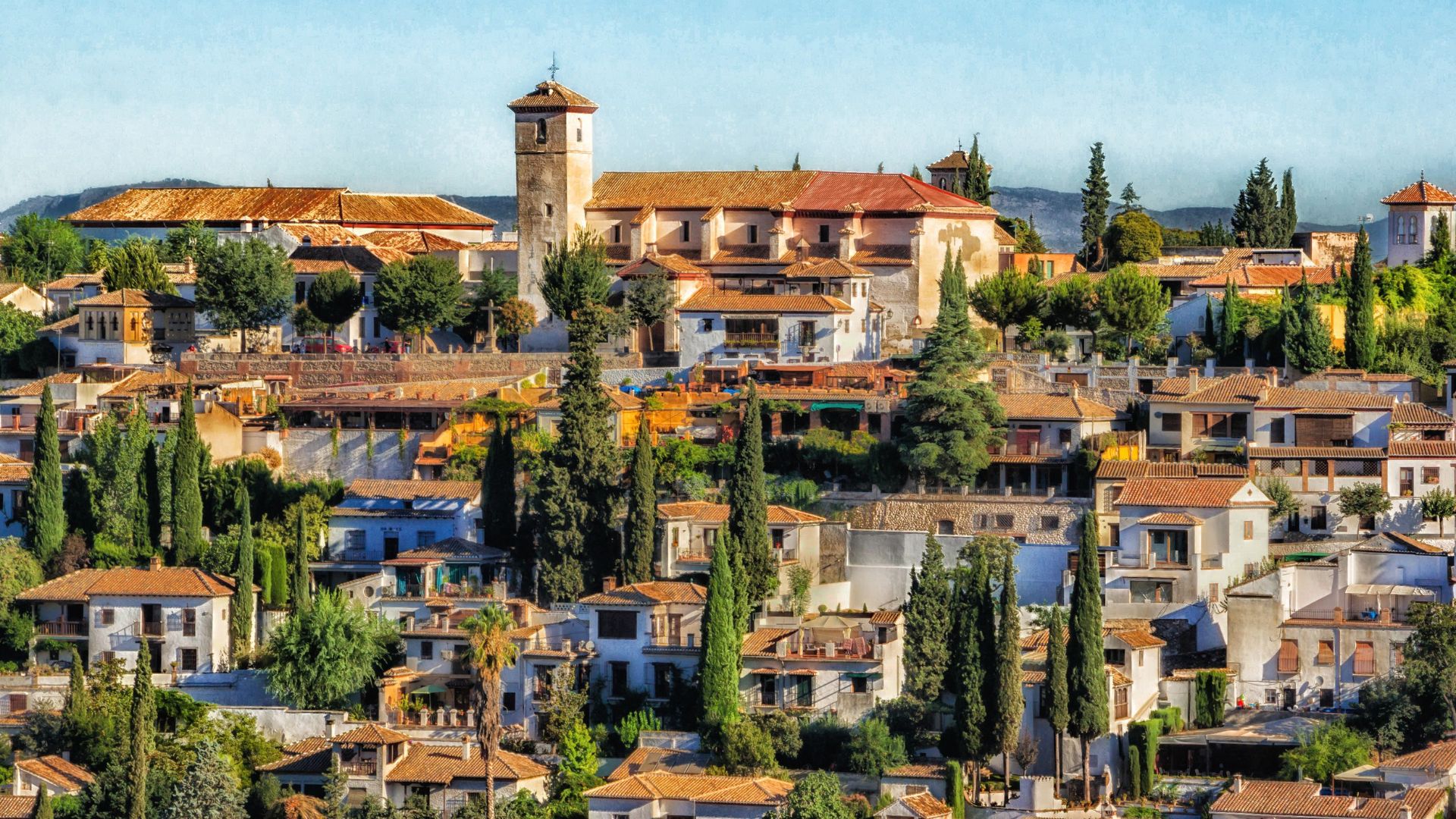 A high-angle view of the historic Albayzín neighborhood in Granada, Spain, featuring traditional white houses with red-tiled roofs nestled on a hillside, surrounded by lush green trees, with the majestic Alhambra Palace and its iconic tower visible in the background under a clear sky.