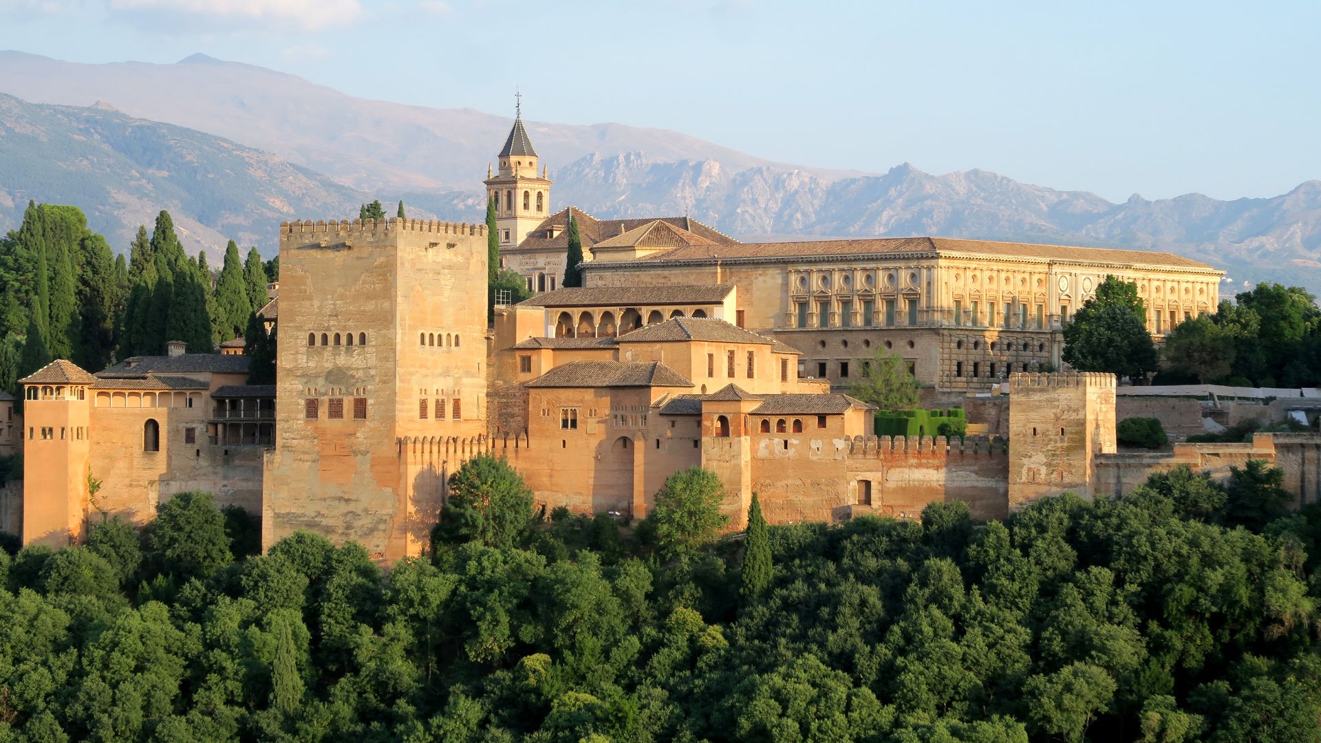 A panoramic view of the historic Alhambra Palace complex, featuring its distinctive towers and walled structures, set atop a hill surrounded by lush green trees, with mountains visible in the background under a clear sky.