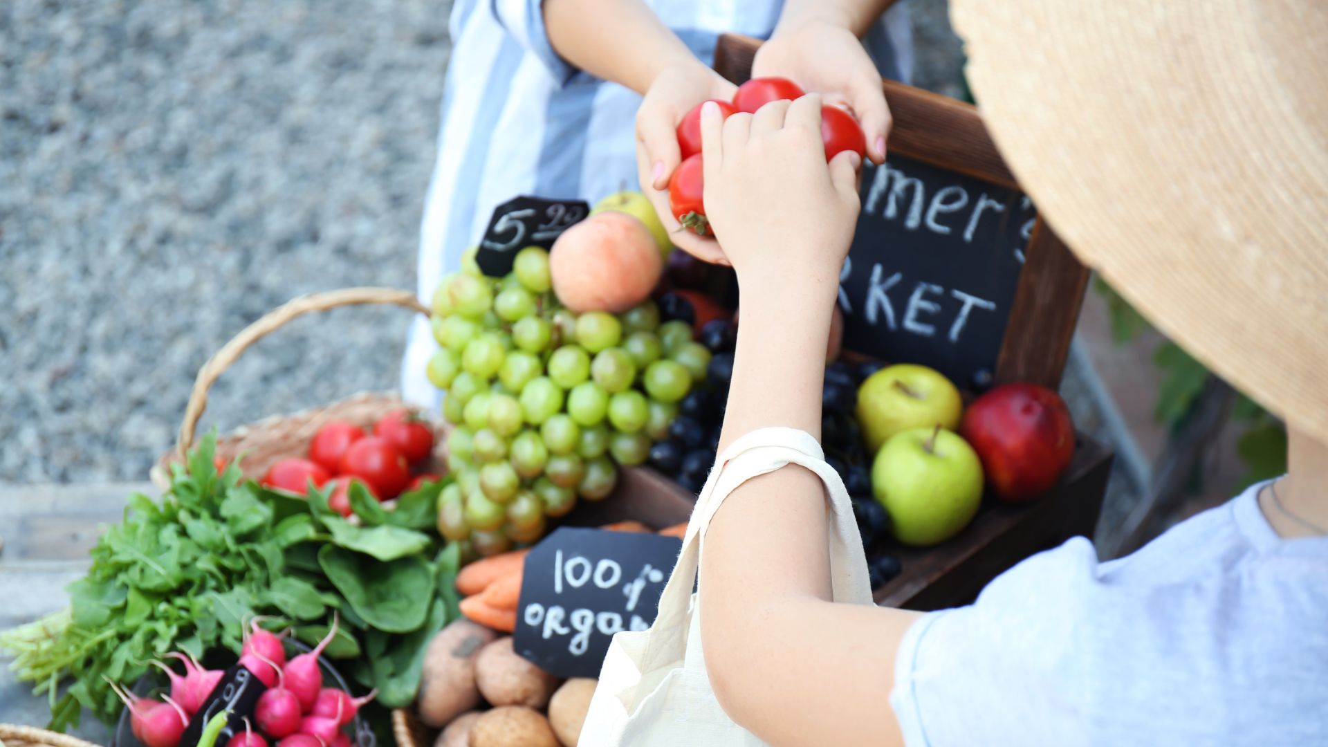A person in a straw hat receives a bunch of red tomatoes from another person at a vibrant outdoor farmers' market stall filled with fresh produce like grapes, apples, radishes, and leafy greens.