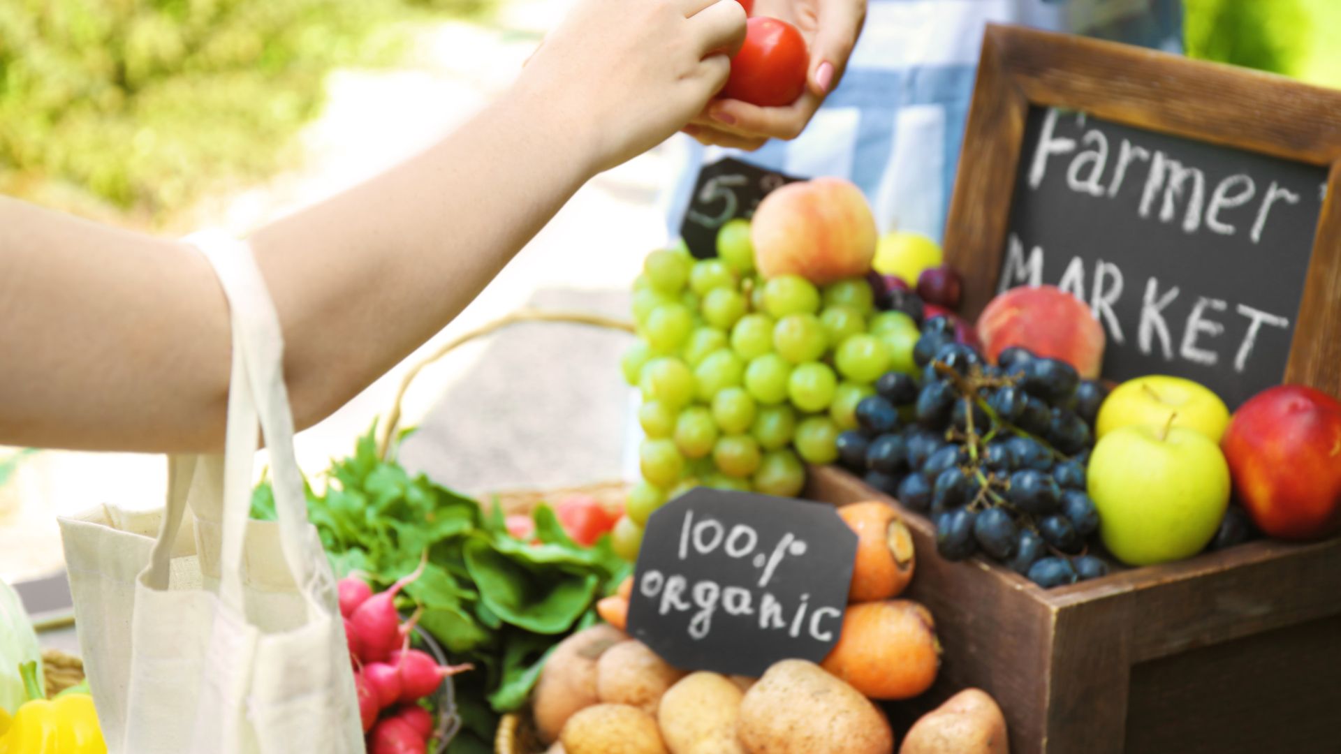A person in a straw hat receives a bunch of red tomatoes from another person at a vibrant outdoor farmers' market stall filled with fresh produce like grapes, apples, radishes, and leafy greens.