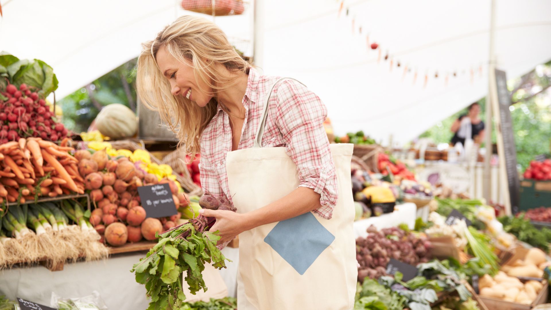 The image shows a woman shopping at what appears to be a farmers market, characterized by stalls laden with fresh produce like fruits and vegetables, and an open-air setting with tents, which are common features of farmers markets globally, including those in the United States. 