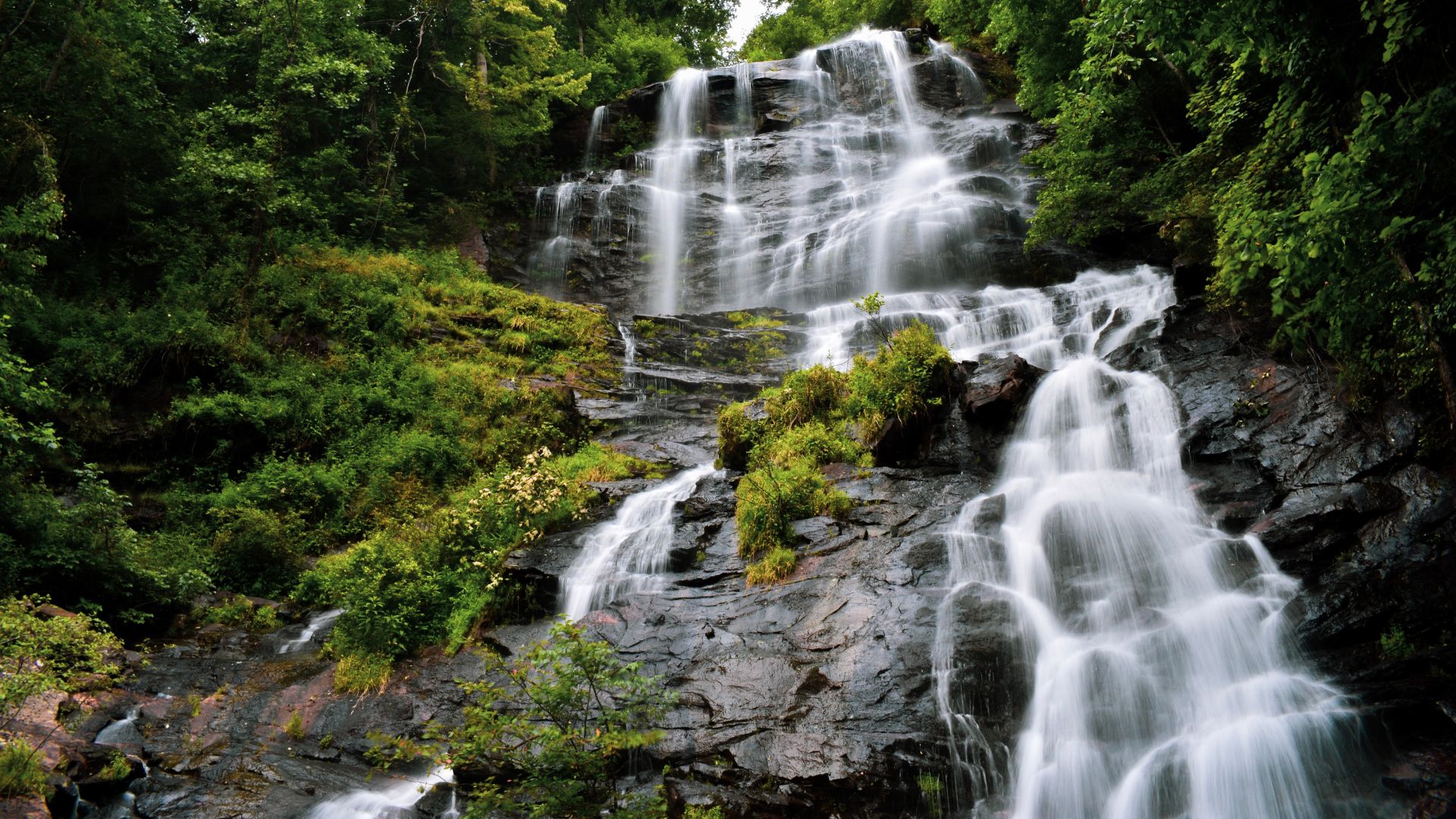 A majestic, multi-tiered waterfall cascades down a rocky, verdant mountainside, surrounded by lush green trees and foliage.