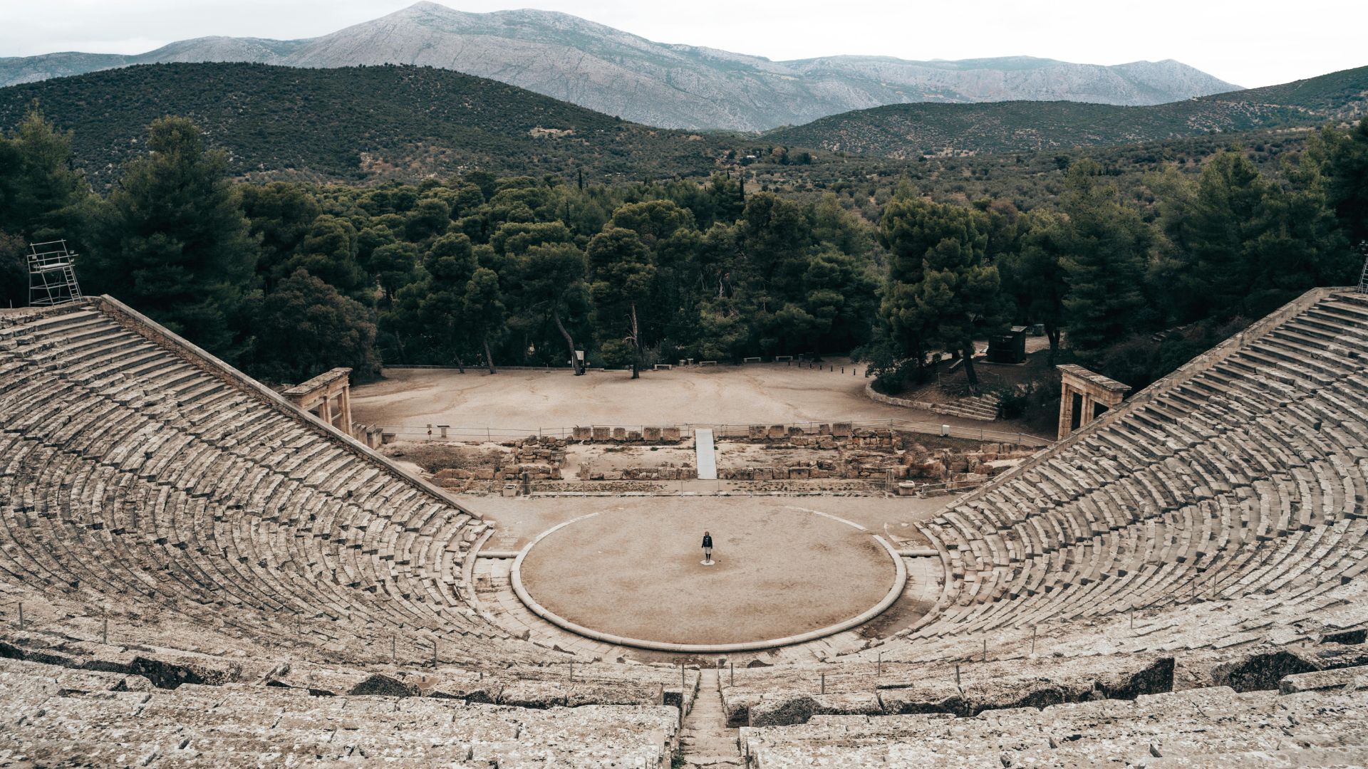 A high-angle view of the large, well-preserved Ancient Theatre of Epidaurus, with its stone seating tiers curving around a central circular orchestra, surrounded by green trees and distant mountains under a cloudy sky.