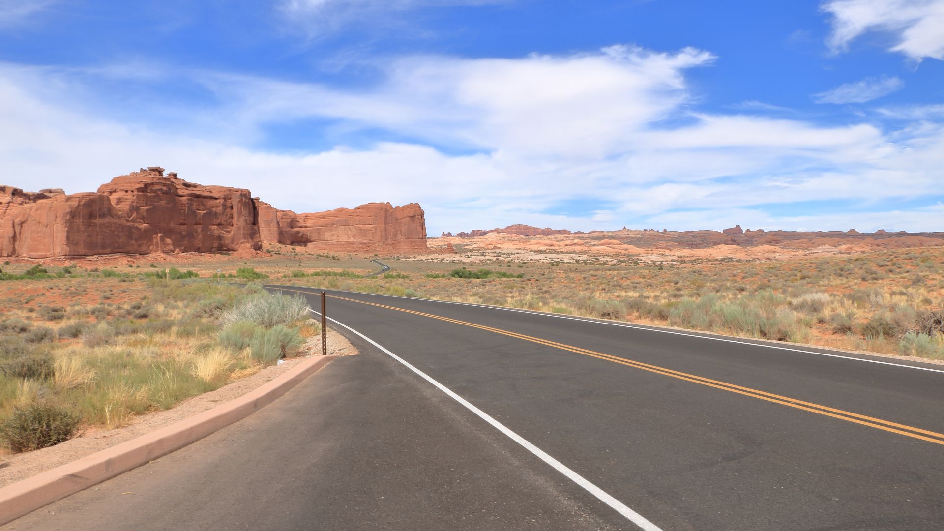A two-lane paved road stretches through a desert landscape with red rock formations under a blue sky with scattered clouds. The road has a white solid line on the right edge and double yellow lines in the center.