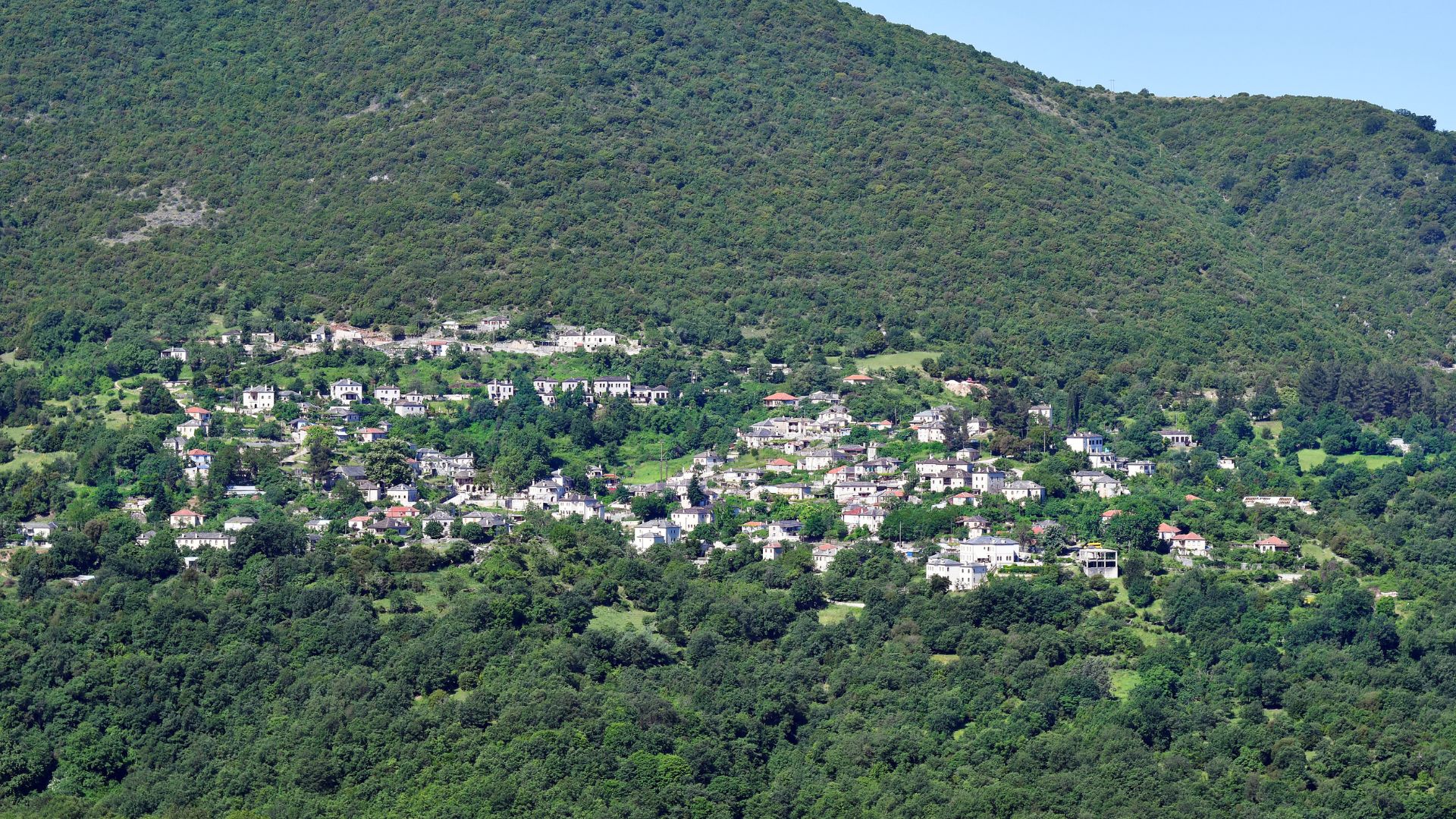 A panoramic view of the traditional village of Aristi nestled amidst a lush green, forested hillside in Greece.