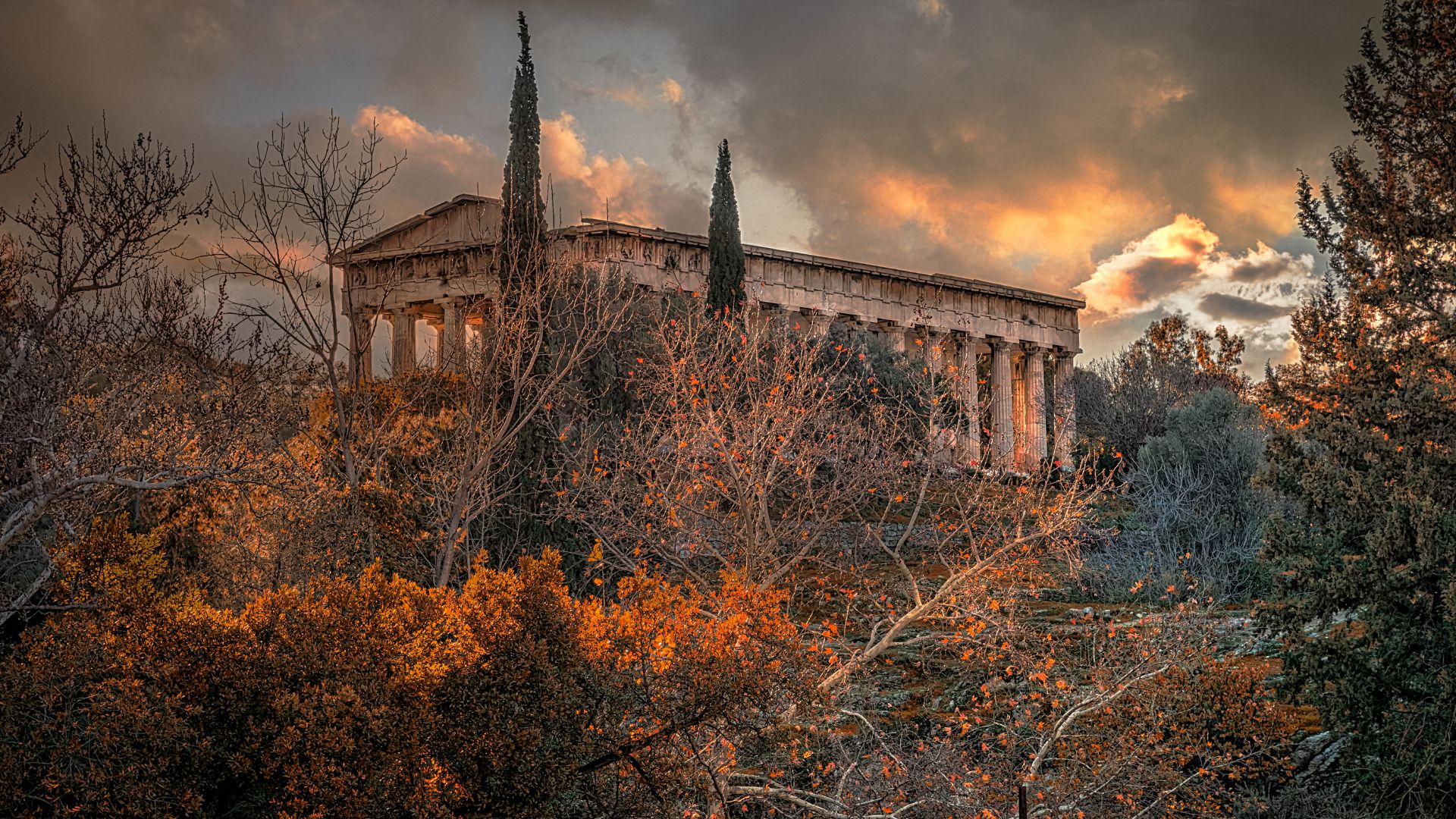 A wide shot of the ancient Temple of Hephaestus in Athens, Greece, seen amidst trees with autumn foliage, under a dramatic, cloudy sunset sky.