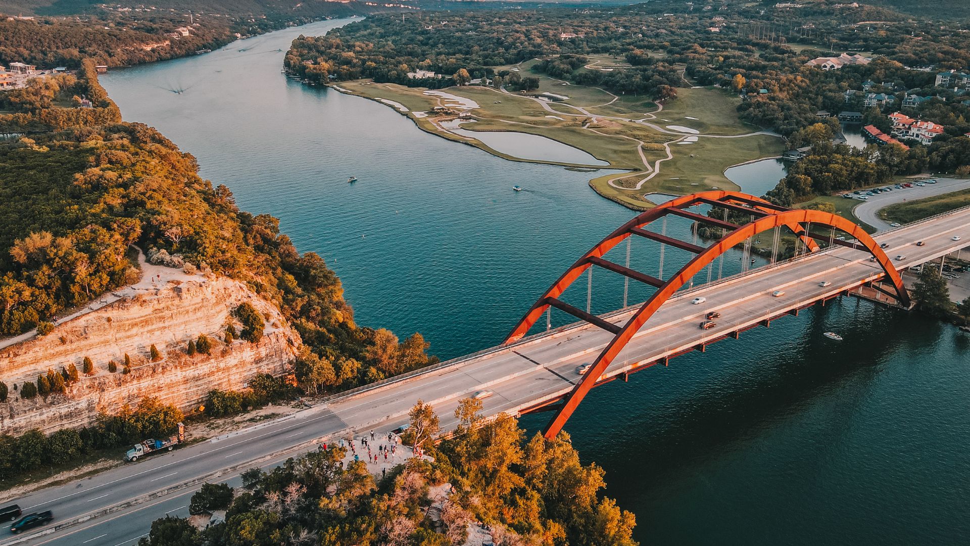 An aerial view of the Pennybacker Bridge, a distinctive steel through-arch bridge with a reddish-brown hue, crossing a wide, blue body of water (Lake Austin). Lush green hills and trees line both sides of the river, with a road leading up to and across the bridge. The setting sun casts warm light on the landscape.