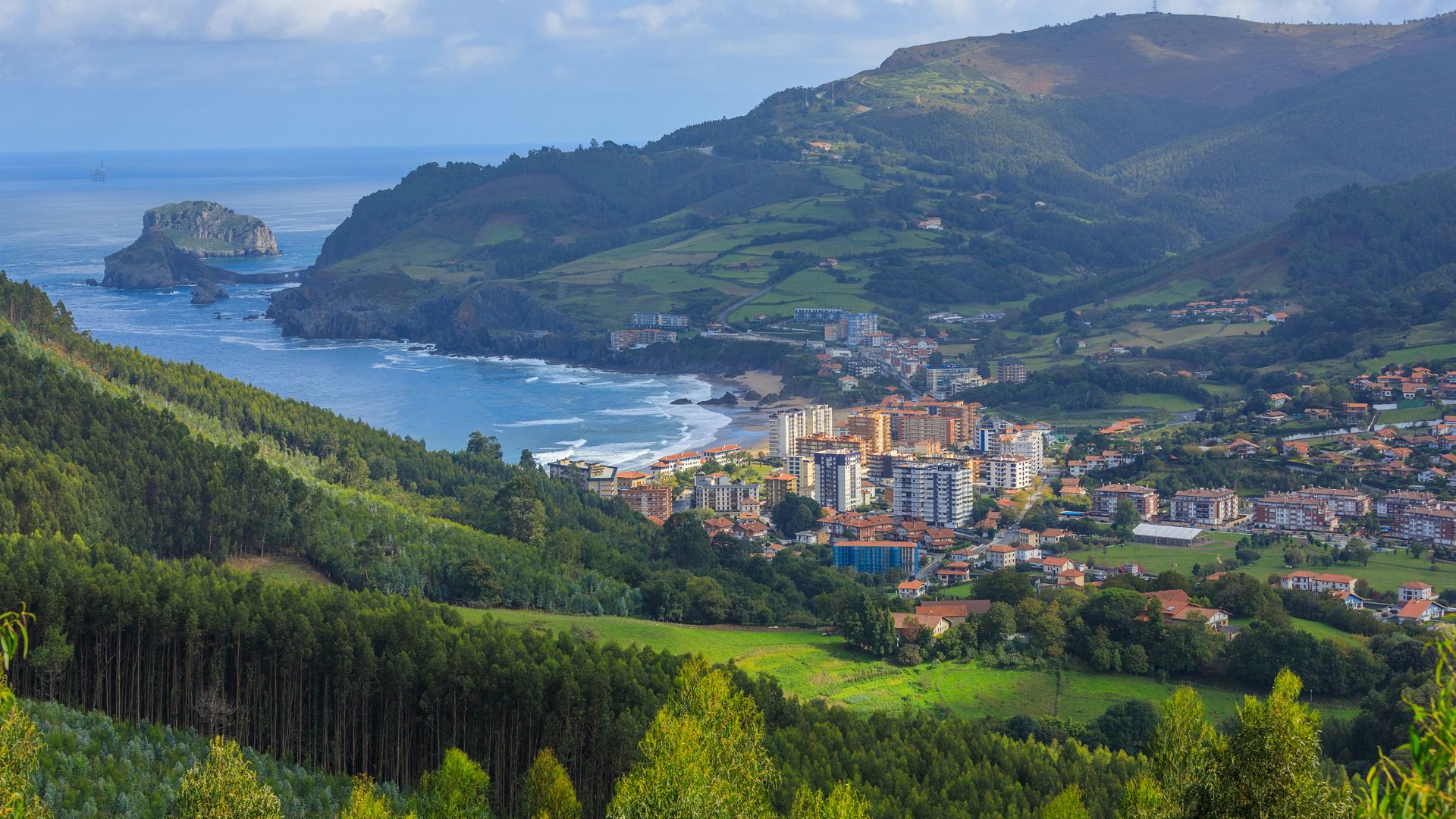 A panoramic view of a coastal town nestled between green, forested mountains and the sea, with a prominent islet and bridge visible in the distance. The town features buildings with orange-tiled roofs and modern structures, all under a bright sky.