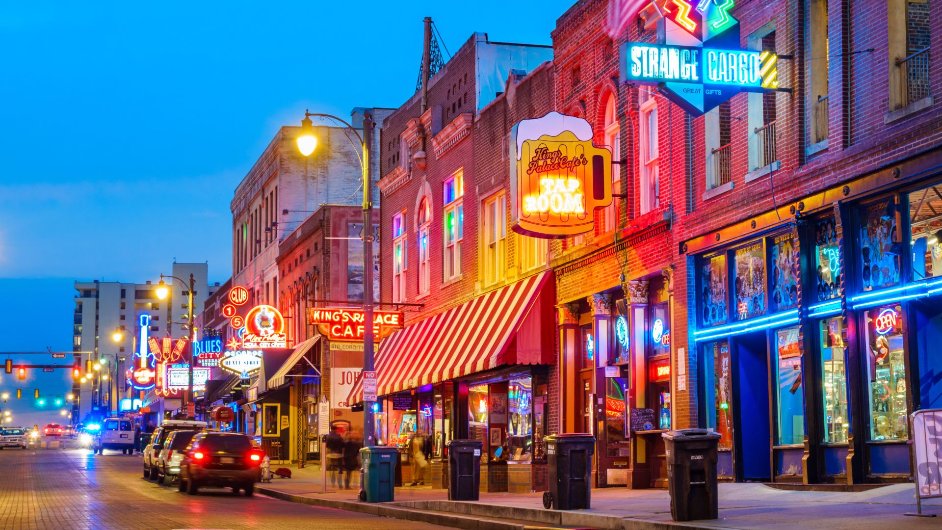 A vibrant night scene of Beale Street in Memphis, Tennessee, featuring historic buildings adorned with colorful neon signs and awnings, with cars and pedestrians on the street. The signs for "STRANGE CARGO" and "KING'S PALACE CAF" are visible, along with a large neon sign shaped like a beer mug advertising "TAP ROOM."