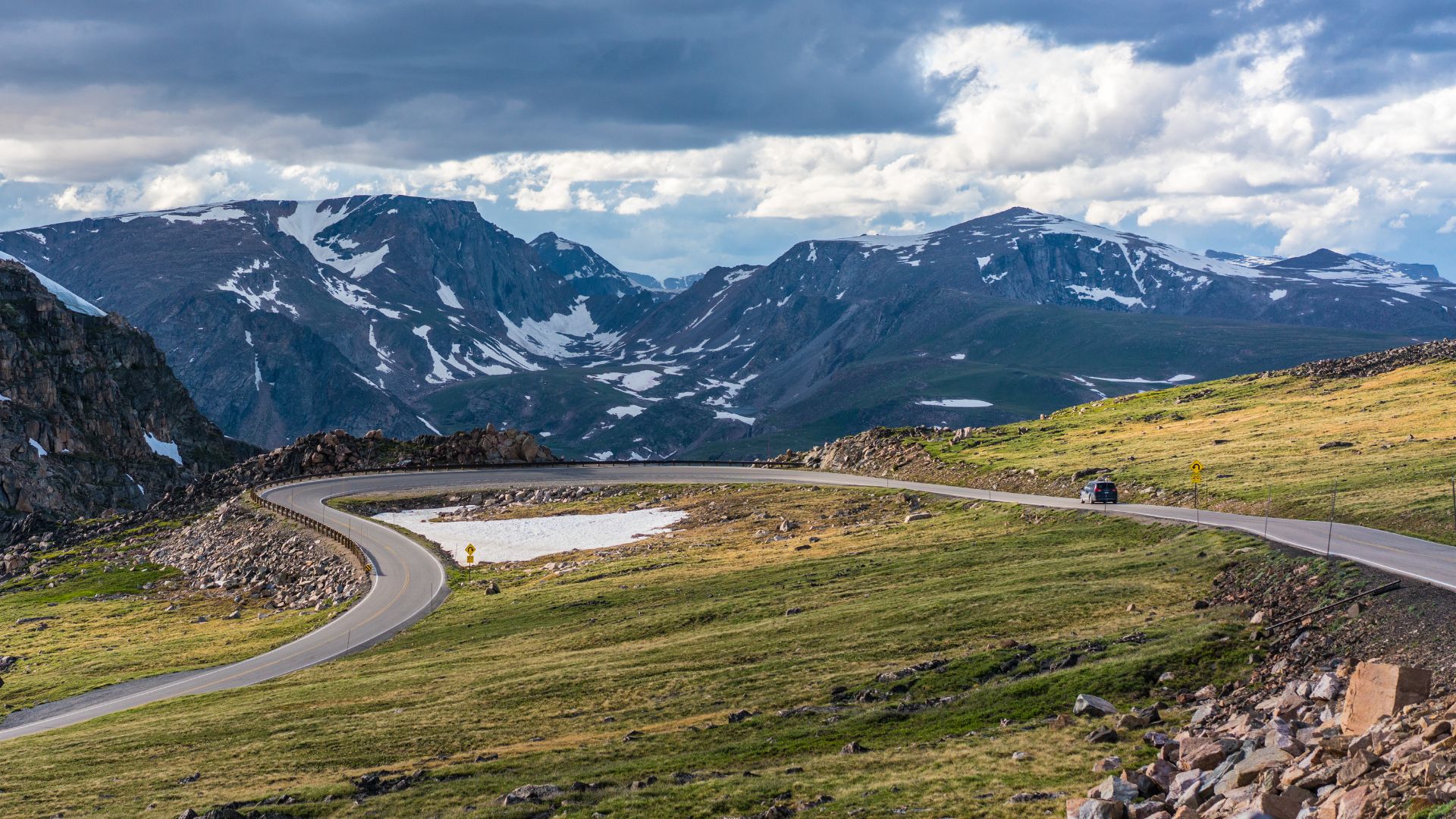  A winding mountain road, part of the Beartooth Highway, curves through a grassy, rocky landscape with snow-capped peaks under a cloudy sky in the background.