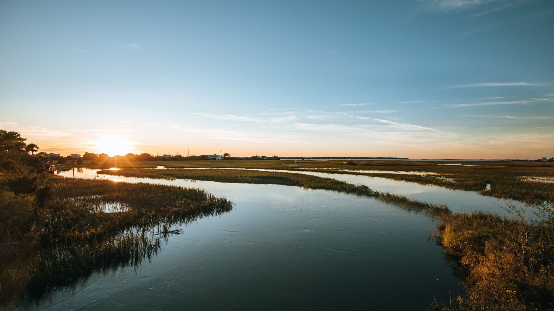 A wide shot of a marshland at sunset, featuring a calm body of water winding through grassy banks under a clear sky with the sun low on the horizon.