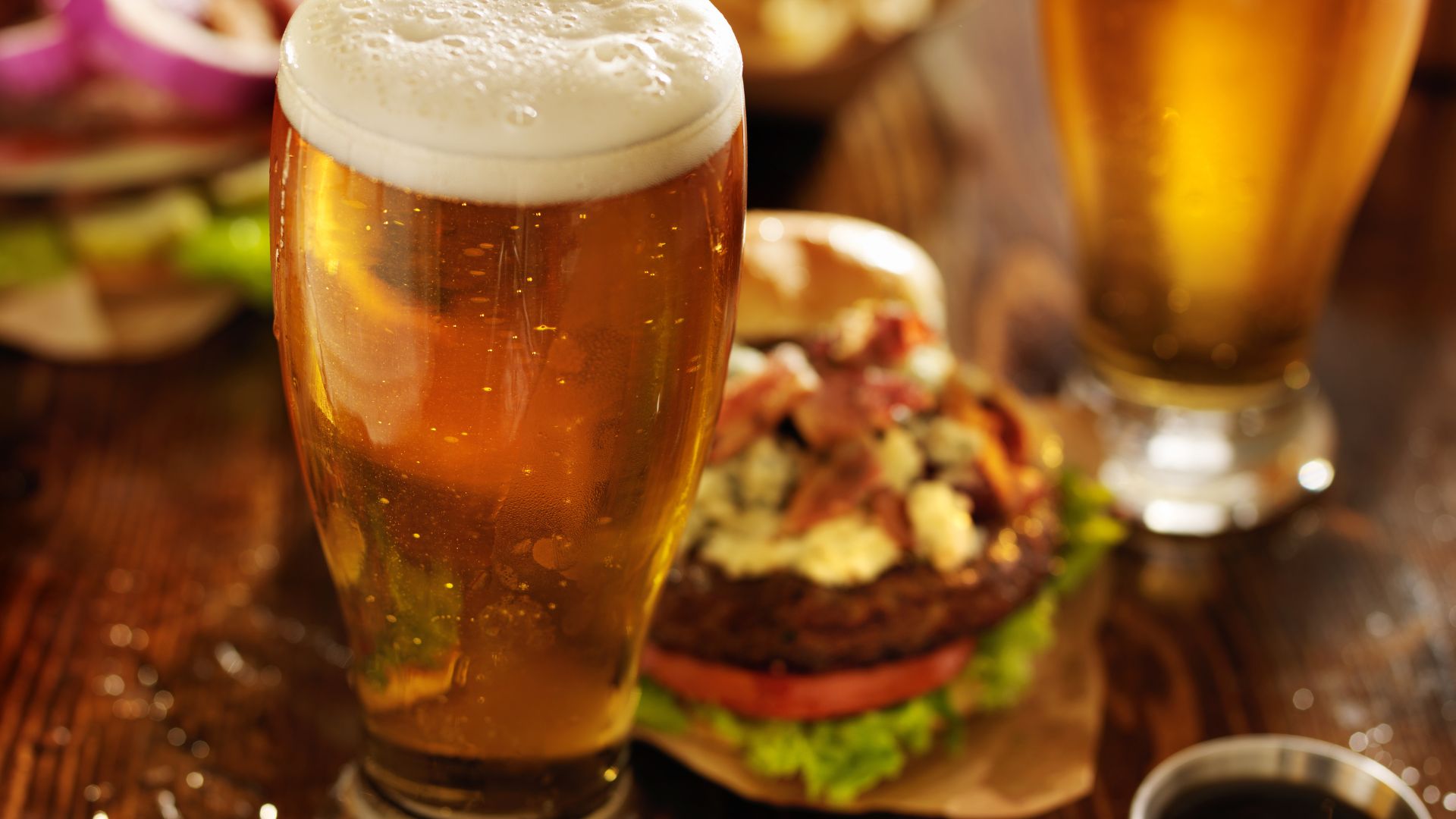 A close-up of a glass of beer with a frothy head, with a burger in the background on a wooden table.
