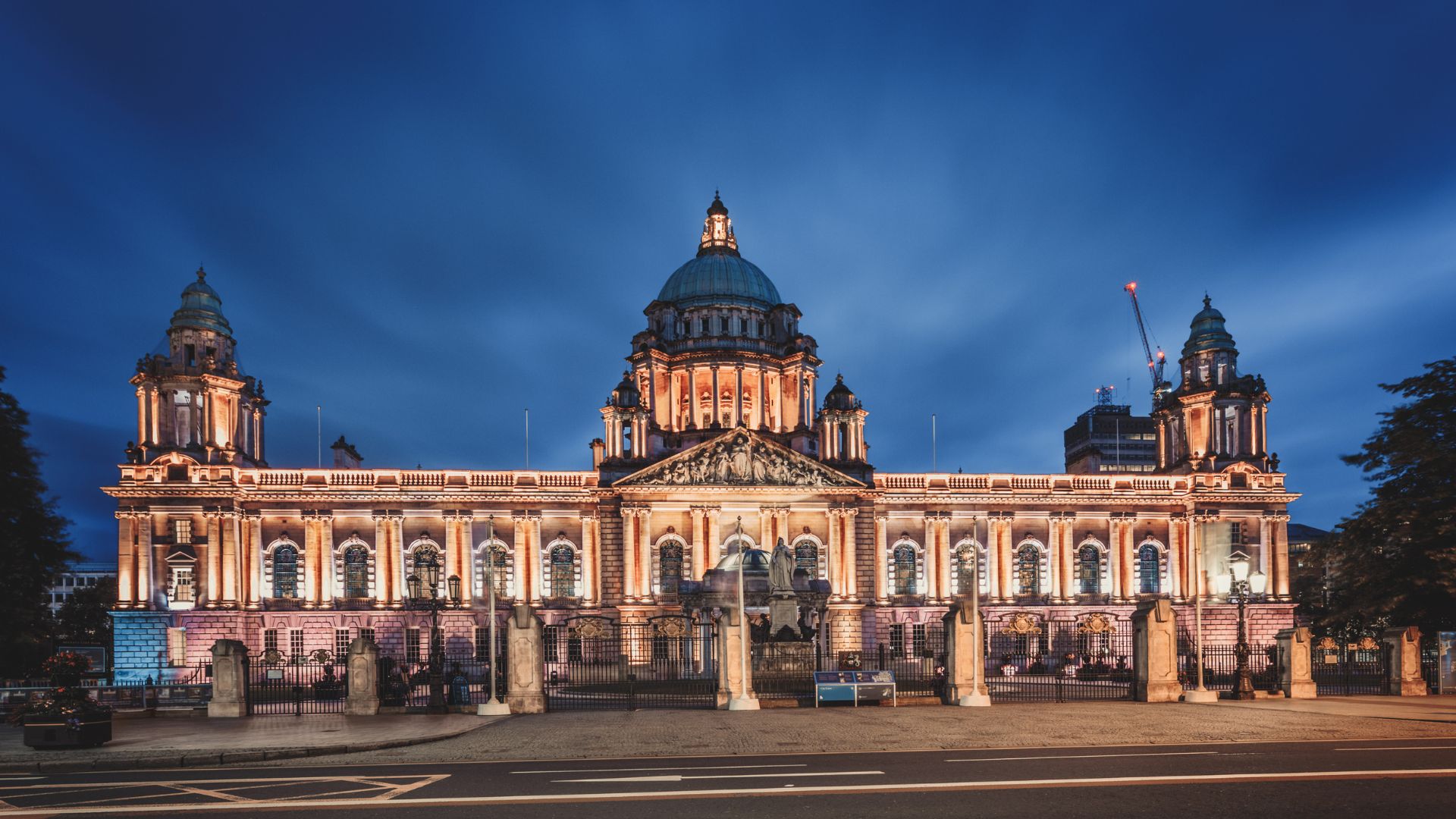Illuminated Belfast City Hall at dusk, a grand civic building with a large central dome and two smaller domes, set against a dark blue sky.