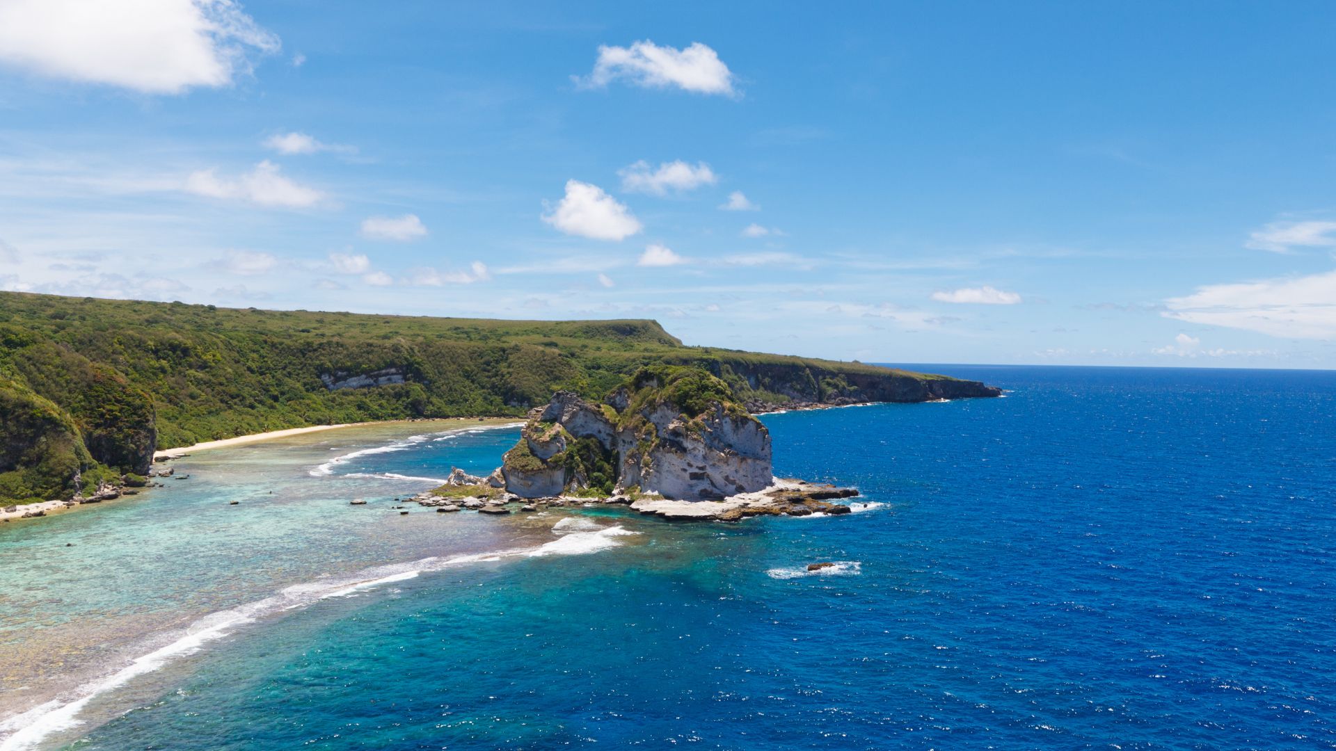 A scenic aerial view of Bird Island, a prominent rock formation in the turquoise waters off the coast of a lush green island with a sandy beach and deep blue ocean under a clear sky with scattered clouds.