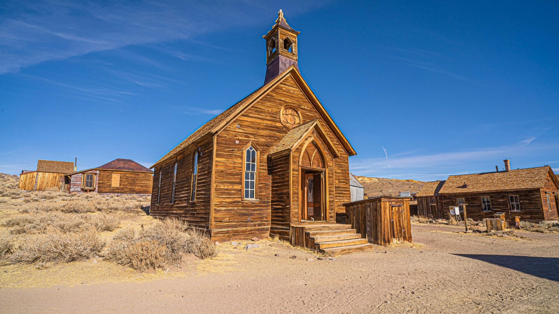 A weathered wooden church with a tall steeple stands in the foreground of a dusty, arid landscape under a clear blue sky, with other abandoned wooden buildings visible in the background, depicting the ghost town of Bodie, California.