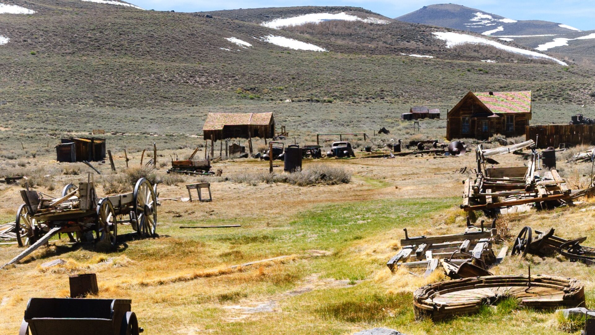 A wide shot of Bodie State Historic Park, a ghost town in California, showing several weathered wooden buildings and scattered remnants of wagons and mining equipment in a dry, grassy landscape with snow-capped hills in the background.