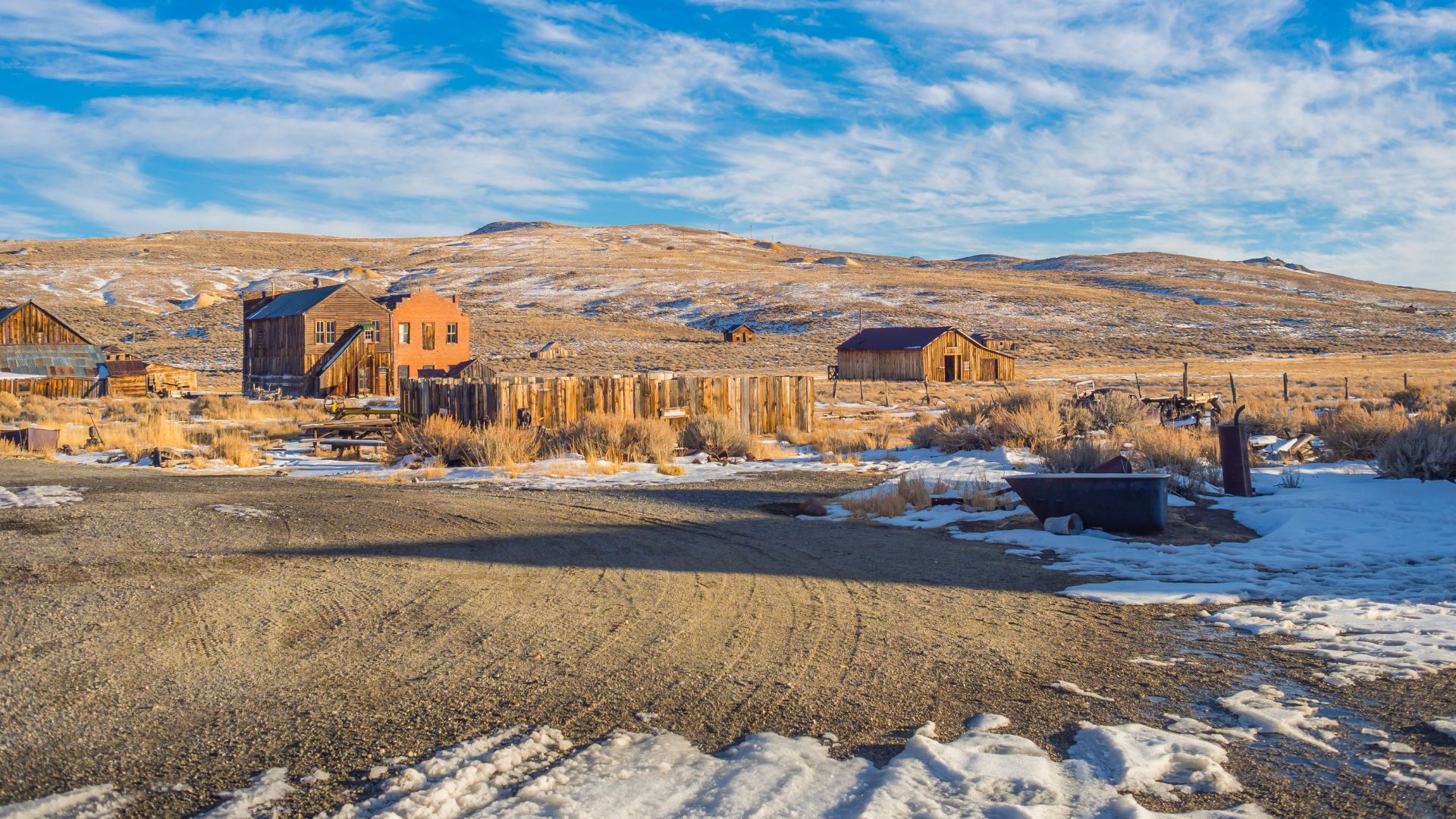 A wide shot of Bodie State Historic Park, a ghost town in California, featuring weathered wooden buildings and structures amidst a snowy, arid landscape under a partly cloudy sky.
