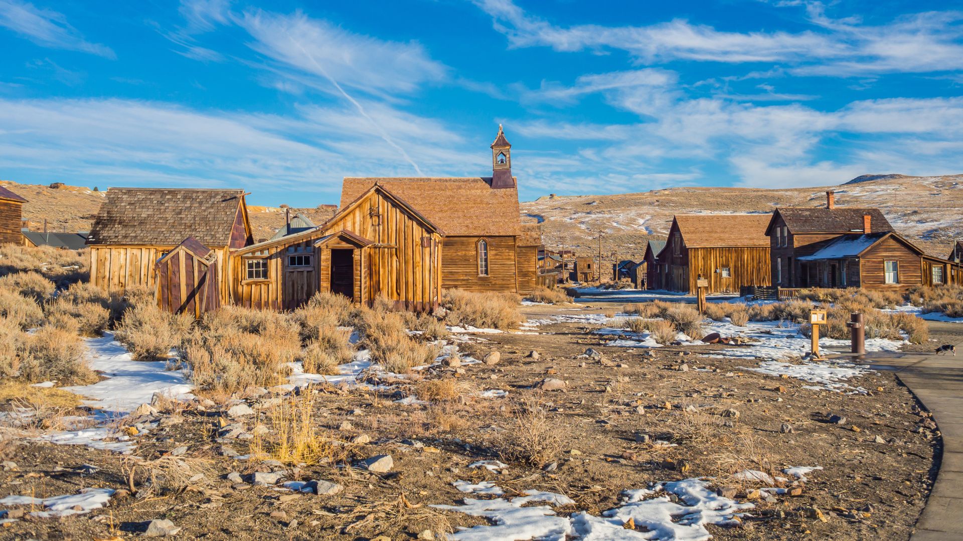 A wide shot of Bodie State Historic Park, a historic gold-mining ghost town in California, featuring several weathered wooden buildings, including a prominent church with a bell tower, scattered across a snow-dusted, arid landscape under a clear blue sky with sparse clouds.