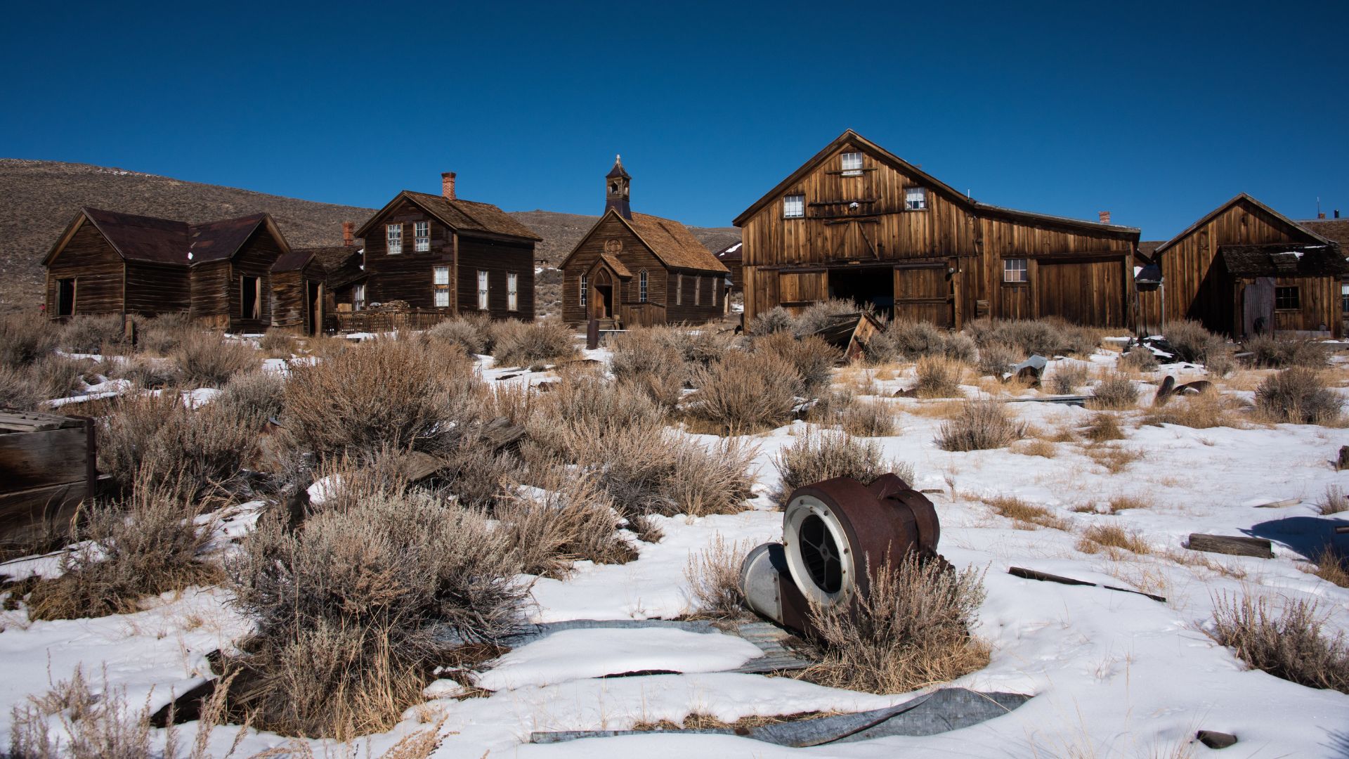 A wide shot of Bodie State Historic Park, a snow-covered ghost town in California, featuring several weathered wooden buildings, including a church and what appear to be homes and a large barn-like structure, under a clear blue sky. In the foreground, dry, bushy vegetation protrudes from the snow, and a large piece of rusted machinery lies partially buried in the snow.