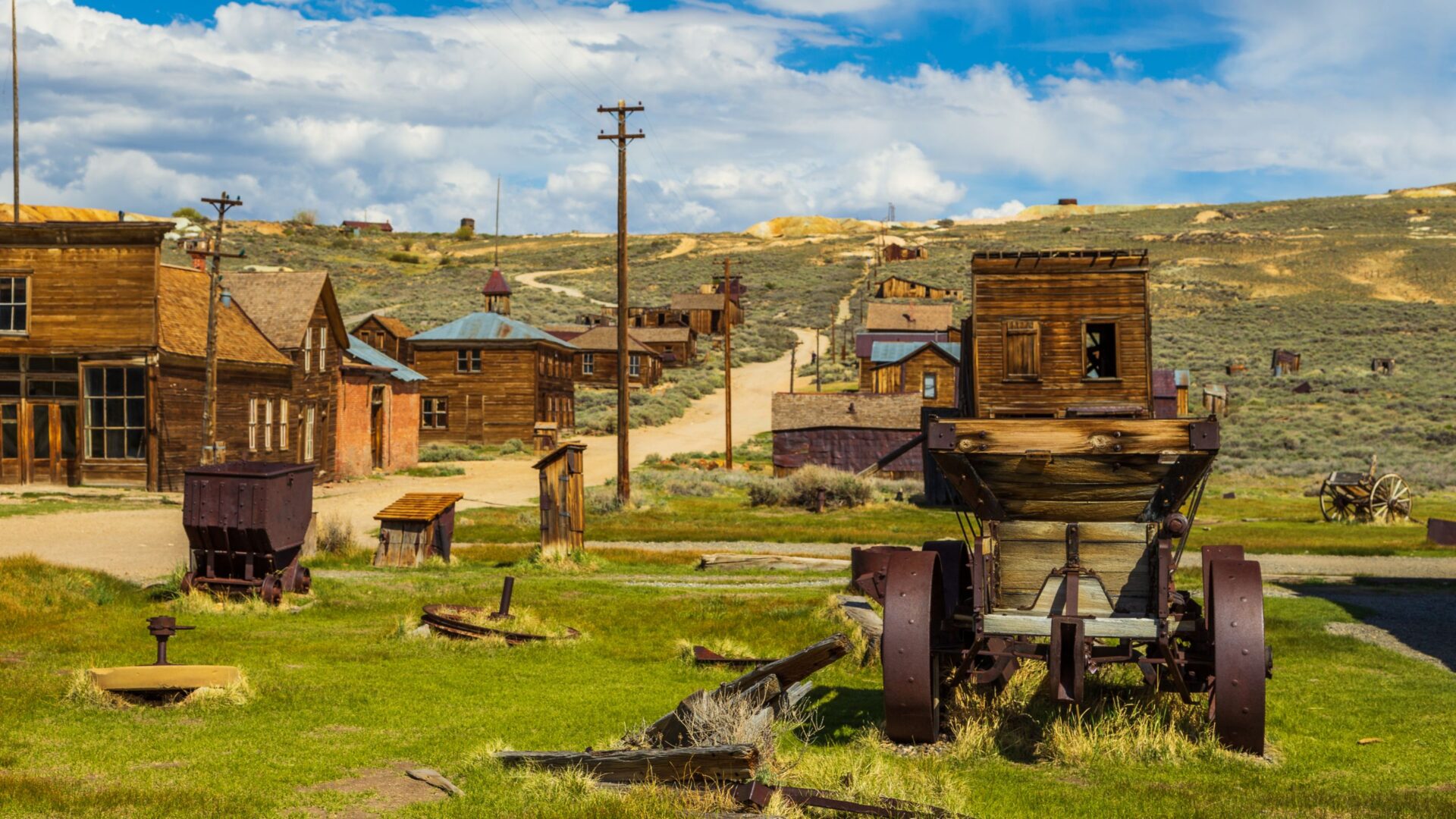 A view of Bodie State Historic Park, a preserved gold-mining ghost town in California, featuring weathered wooden buildings, a dirt road, and an old mining cart in the foreground, all under a partly cloudy sky.