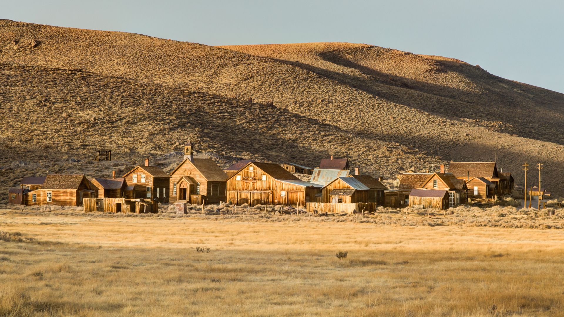 A wide shot of Bodie State Historic Park in California, showing a collection of weathered wooden buildings of a ghost town nestled in a dry, grassy landscape with a large, golden-brown hill rising behind them under a bright sky.