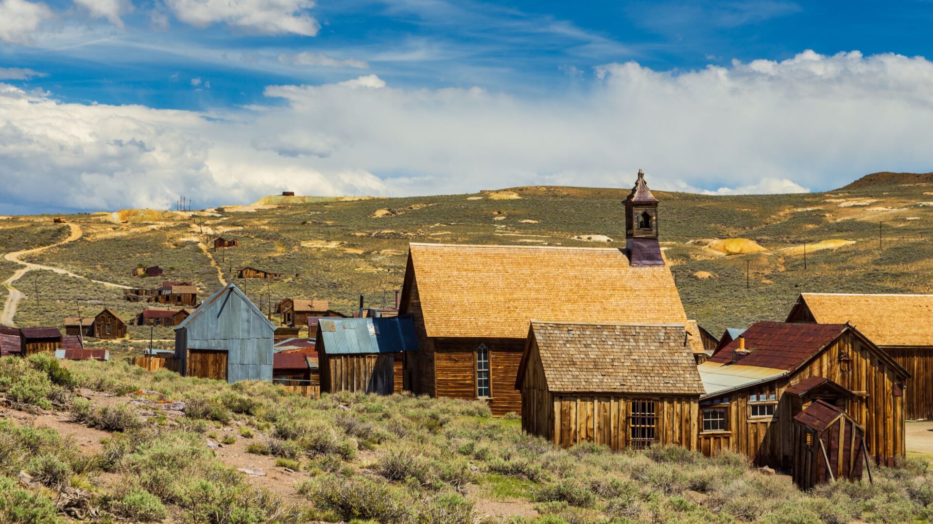 A wide shot of Bodie State Historic Park, a historic California gold-mining ghost town, featuring a prominent wooden church with a bell tower and other weathered wooden buildings scattered across a dry, grassy landscape under a partly cloudy sky.