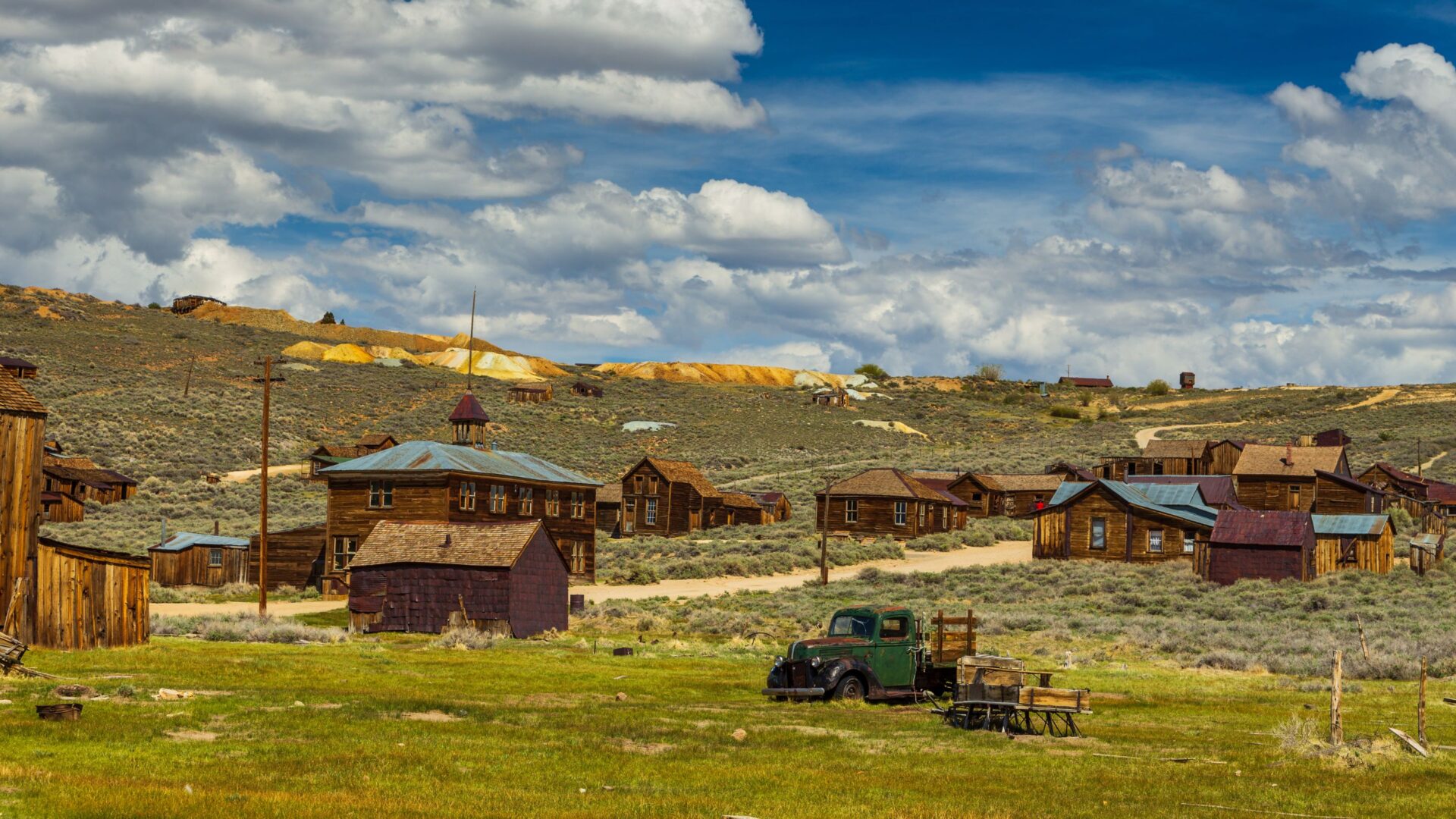 A panoramic view of Bodie State Historic Park, a historic gold-mining ghost town in California, featuring weathered wooden buildings, an old green truck, and sparse vegetation under a cloudy sky with hills in the background.
