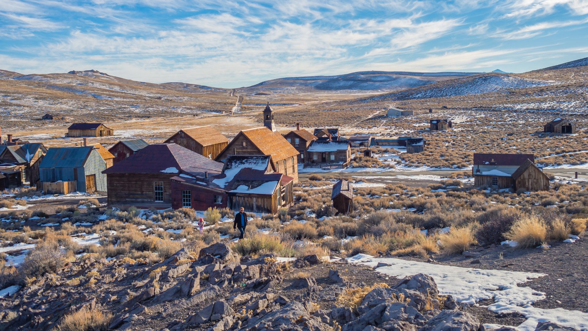 An aerial view of Bodie State Historic Park, a historic gold-mining ghost town in California, featuring numerous weathered wooden buildings scattered across a snow-dusted, arid landscape with rolling hills under a partly cloudy sky.