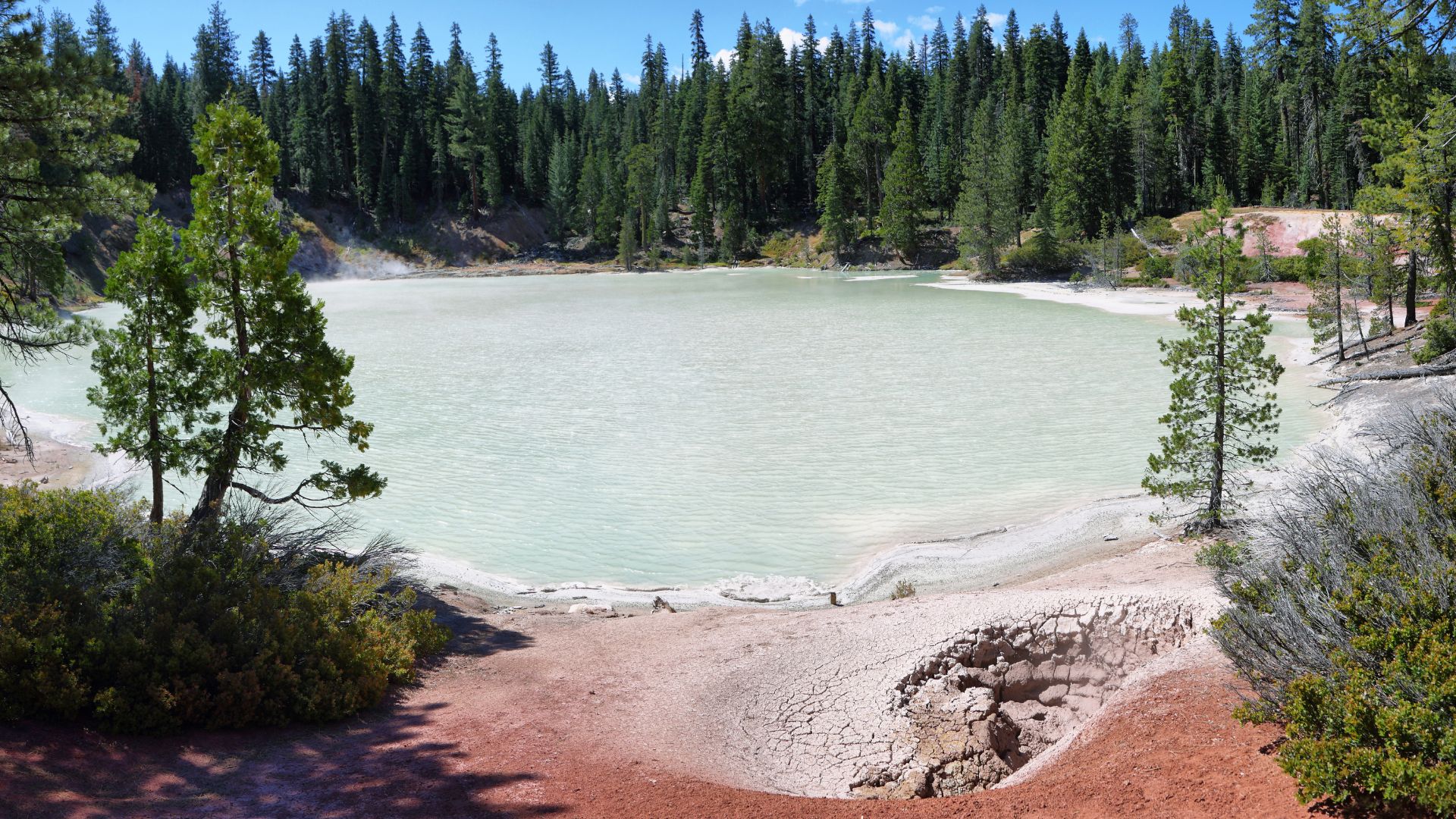 A wide shot of Boiling Springs Lake in Lassen Volcanic National Park, California, featuring a milky blue-green lake with steam rising from its surface, surrounded by dense green pine forests, and backed by distant mountains under a blue sky with scattered white clouds.