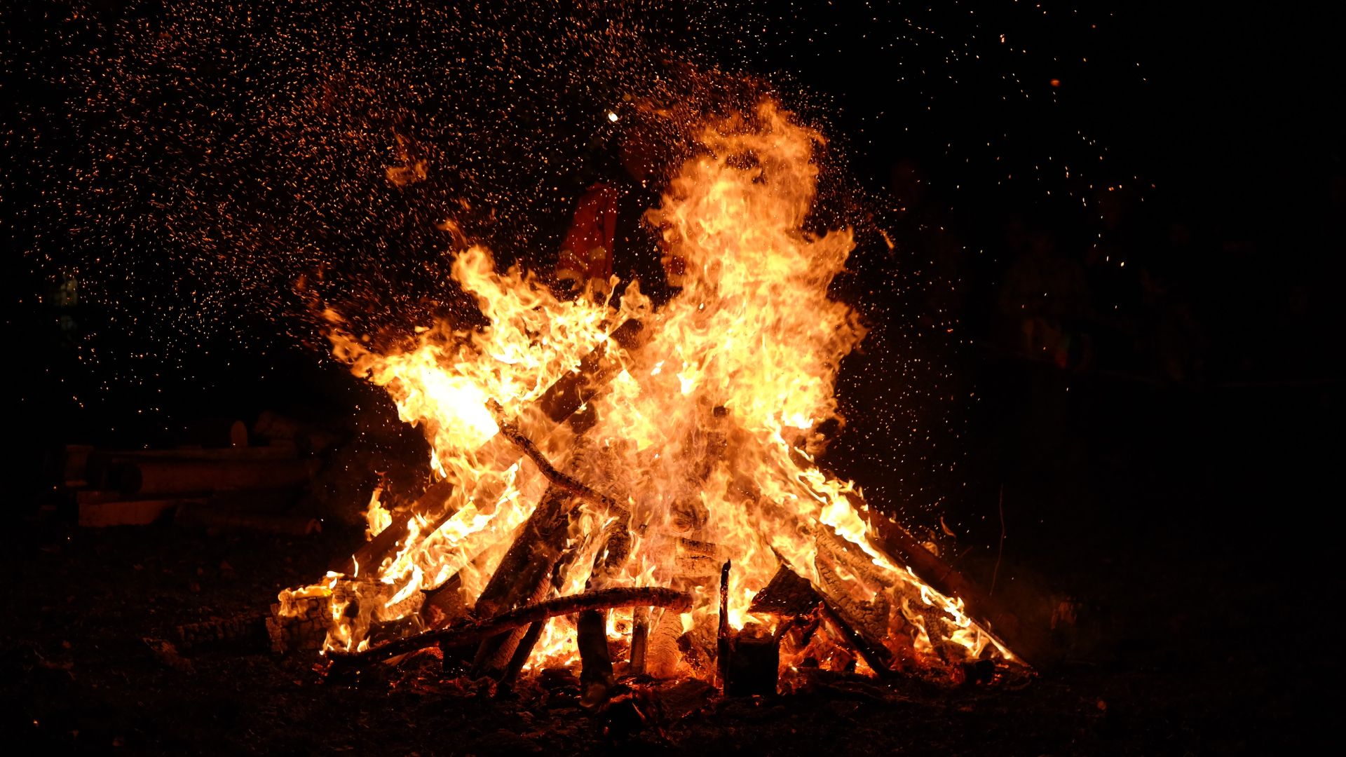 A large, intense bonfire blazing at night, with bright orange and yellow flames engulfing what appears to be a wooden structure or effigy.