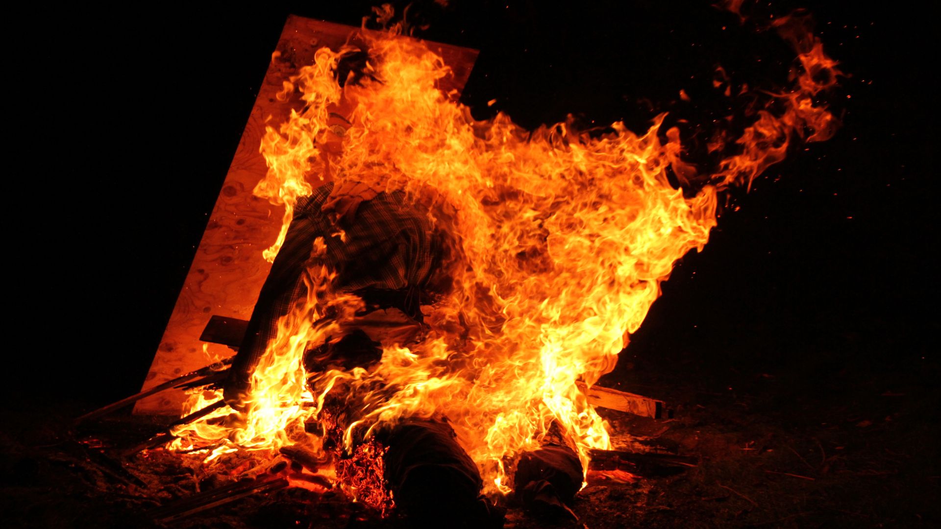 A large, intense bonfire blazing at night, with bright orange and yellow flames engulfing what appears to be a wooden structure or effigy.
