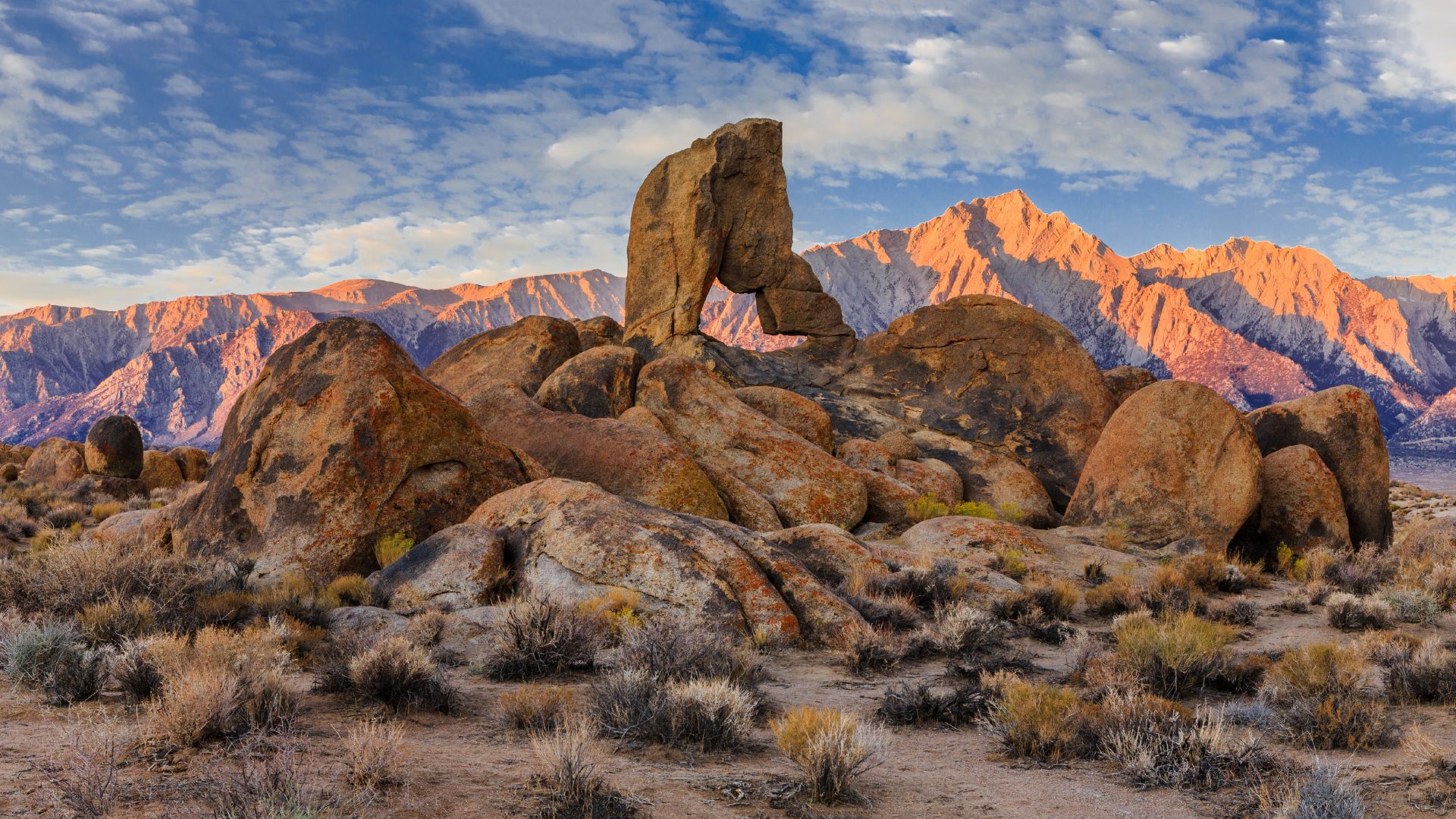 A wide shot of Boot Arch, a natural rock formation resembling a boot, in the Alabama Hills of California, with mountains illuminated by warm light in the background under a partly cloudy sky.