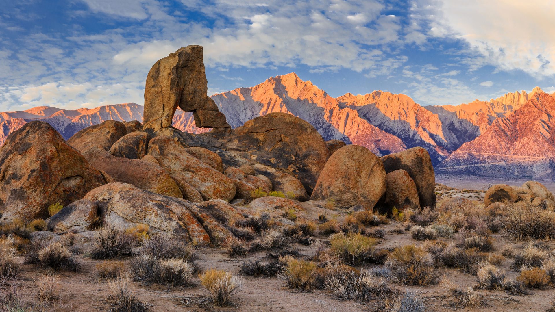 A natural rock arch formation, known as Boot Arch, stands prominently in the foreground of the Alabama Hills, with the sunlit peaks of the Sierra Nevada mountains in the background under a partly cloudy sky.