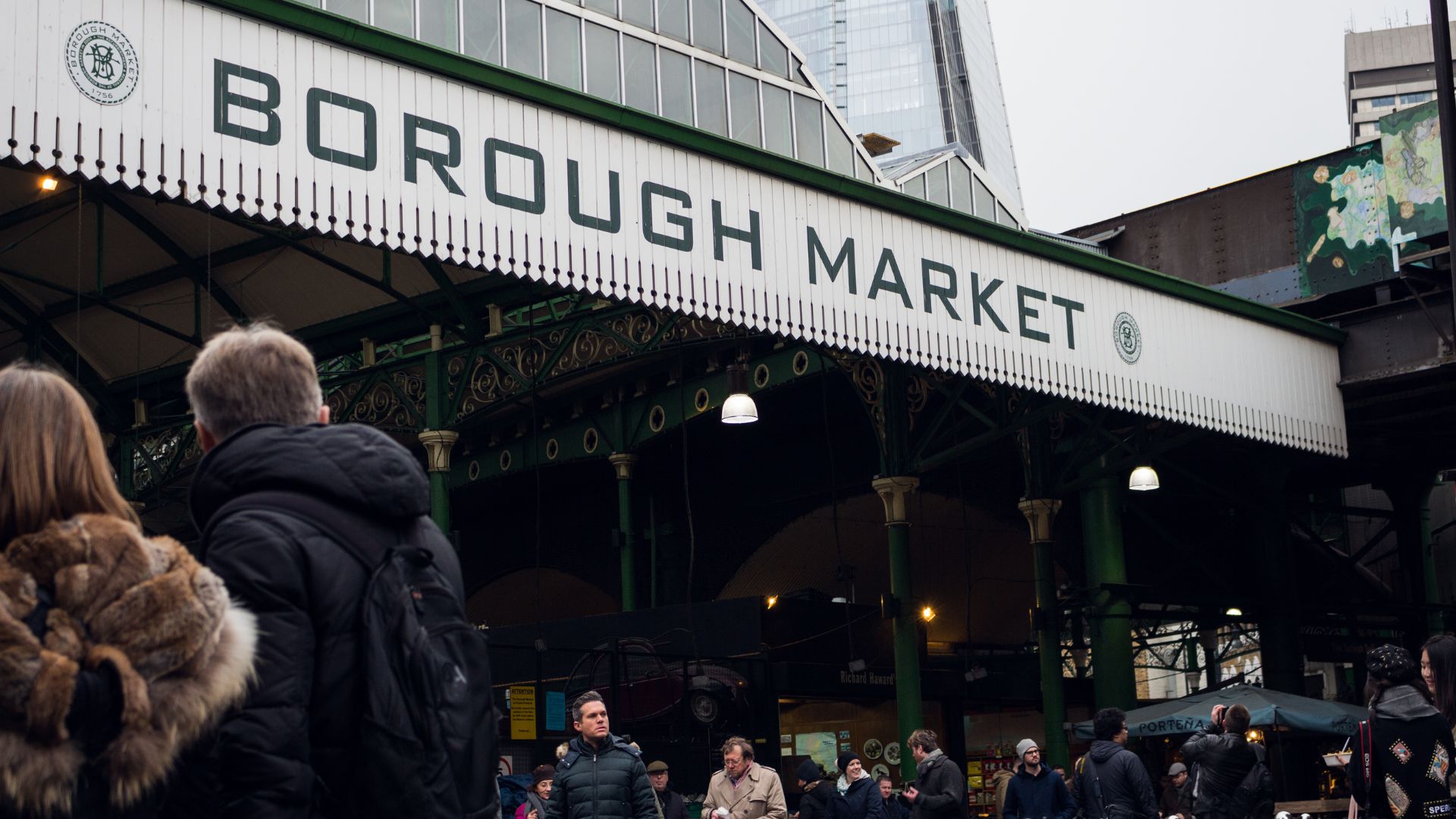 A bustling outdoor market scene with a large sign overhead reading "BOROUGH MARKET," featuring people browsing stalls and the distinctive architecture of the market hall.