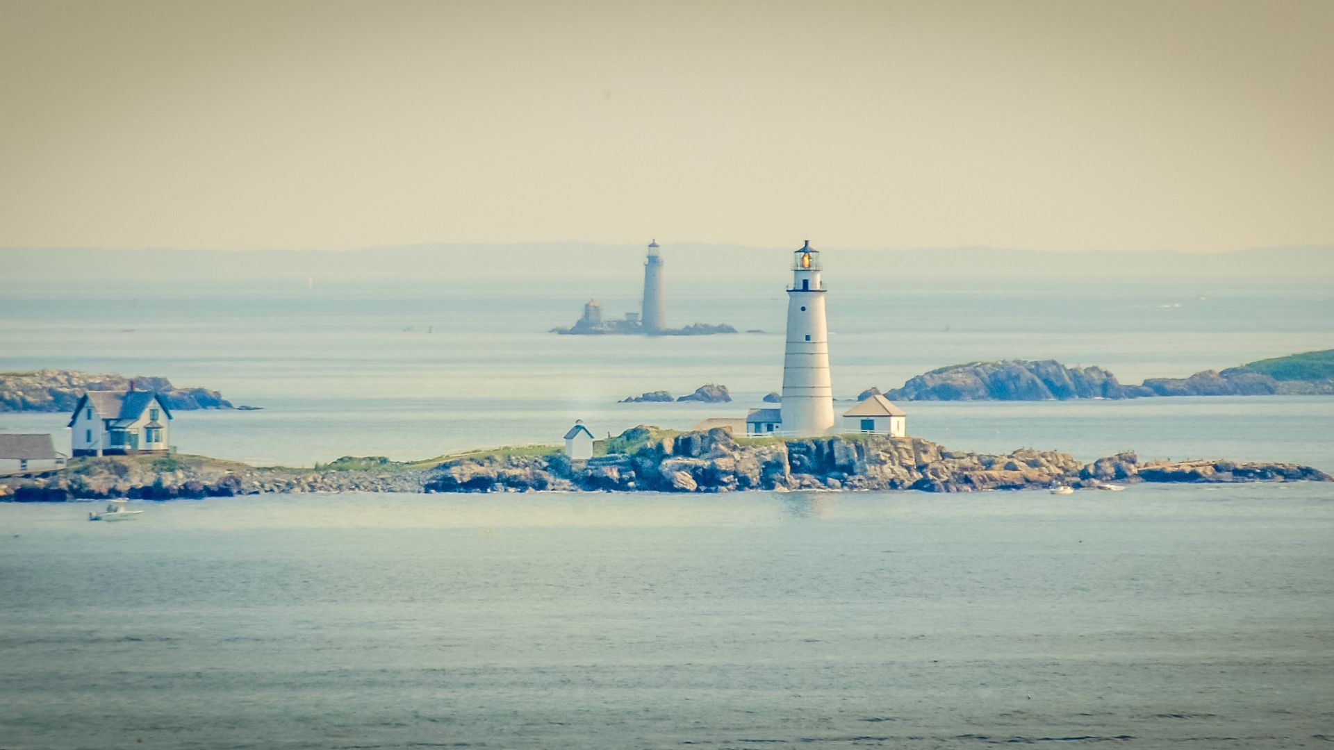 A long-distance shot of Boston Light, a white lighthouse with a red roof, situated on a rocky island in the middle of a calm body of water. Another, more distant lighthouse can be seen further out in the hazy background, and a small house is visible on the left side of the main island.