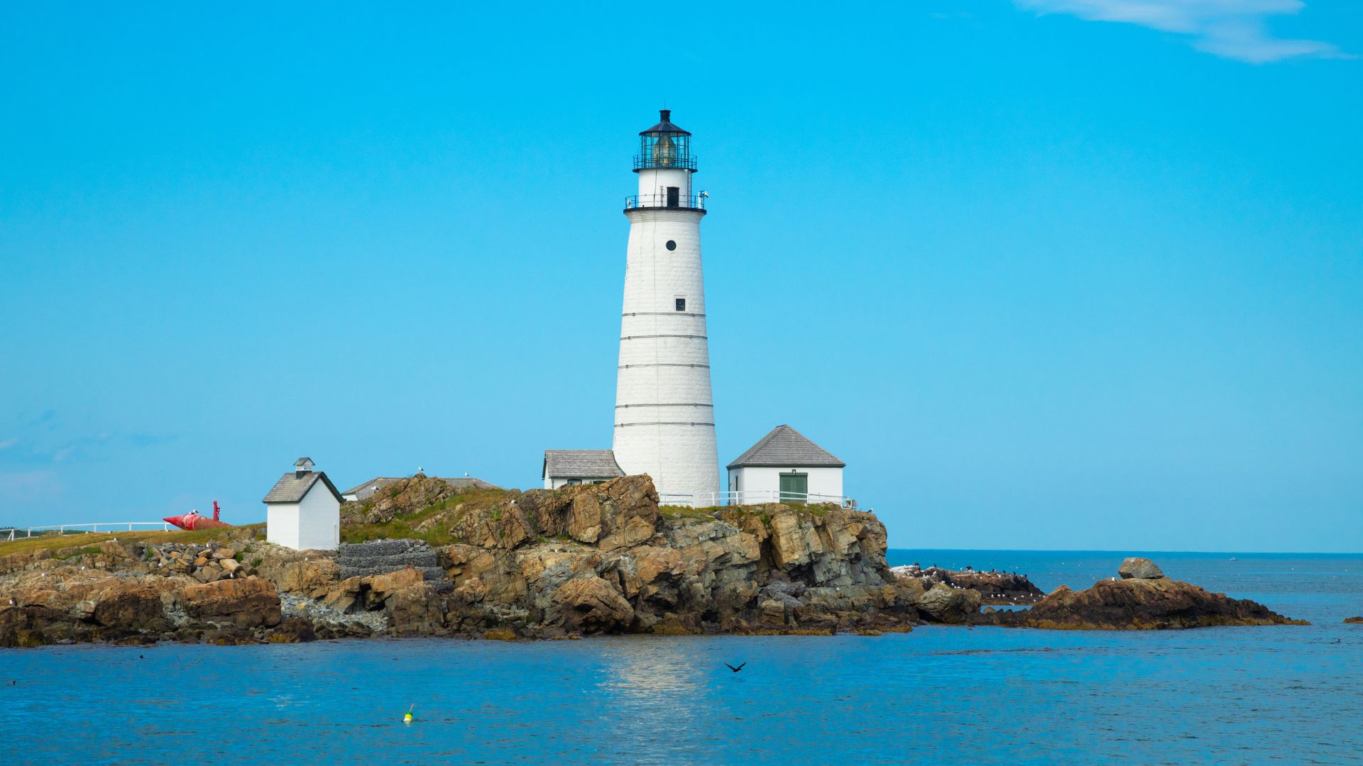 A tall, white lighthouse with a black top stands on a rocky island surrounded by blue water under a clear blue sky, with smaller white buildings nestled near its base.