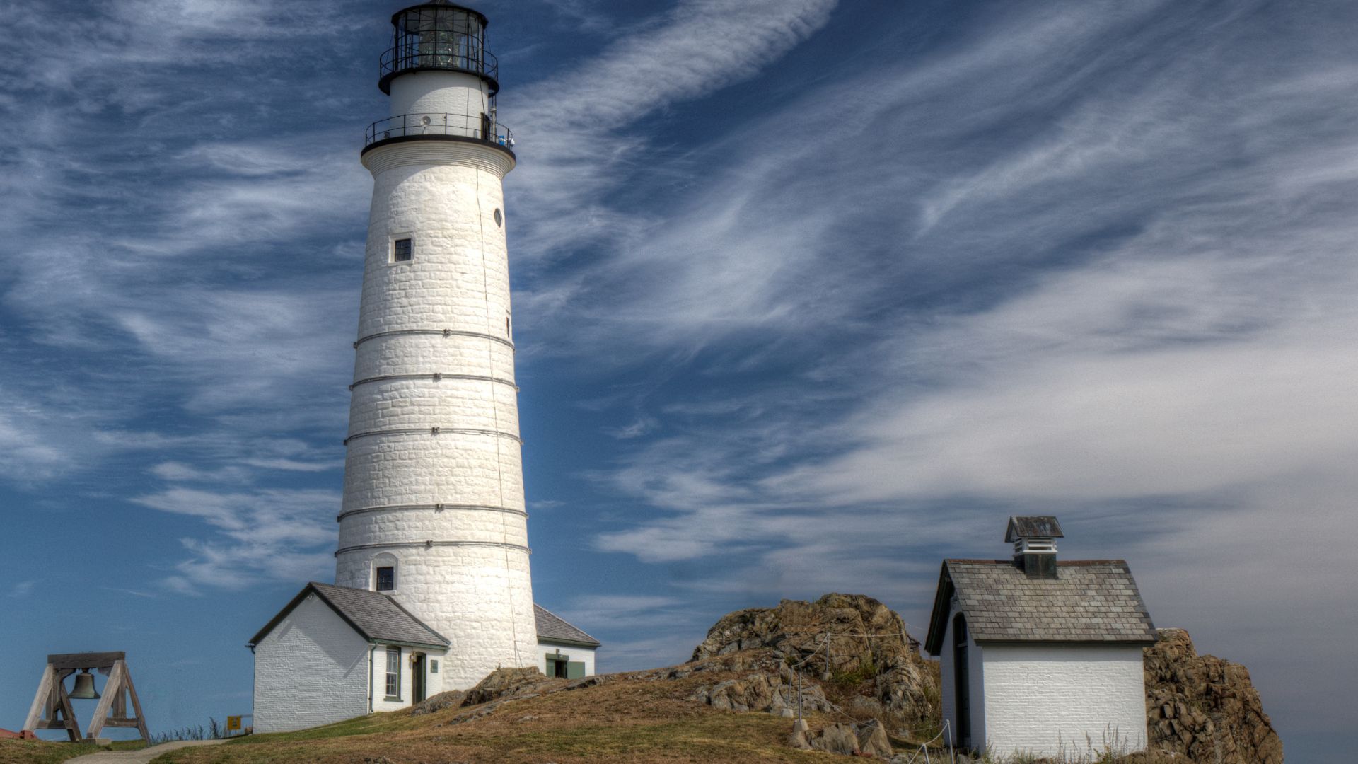 A tall, white lighthouse, Boston Light, stands prominently on a rocky island under a dramatic sky with wispy clouds. Several smaller white buildings are visible around the base of the lighthouse and among the rocks, with a bell structure on the left. 