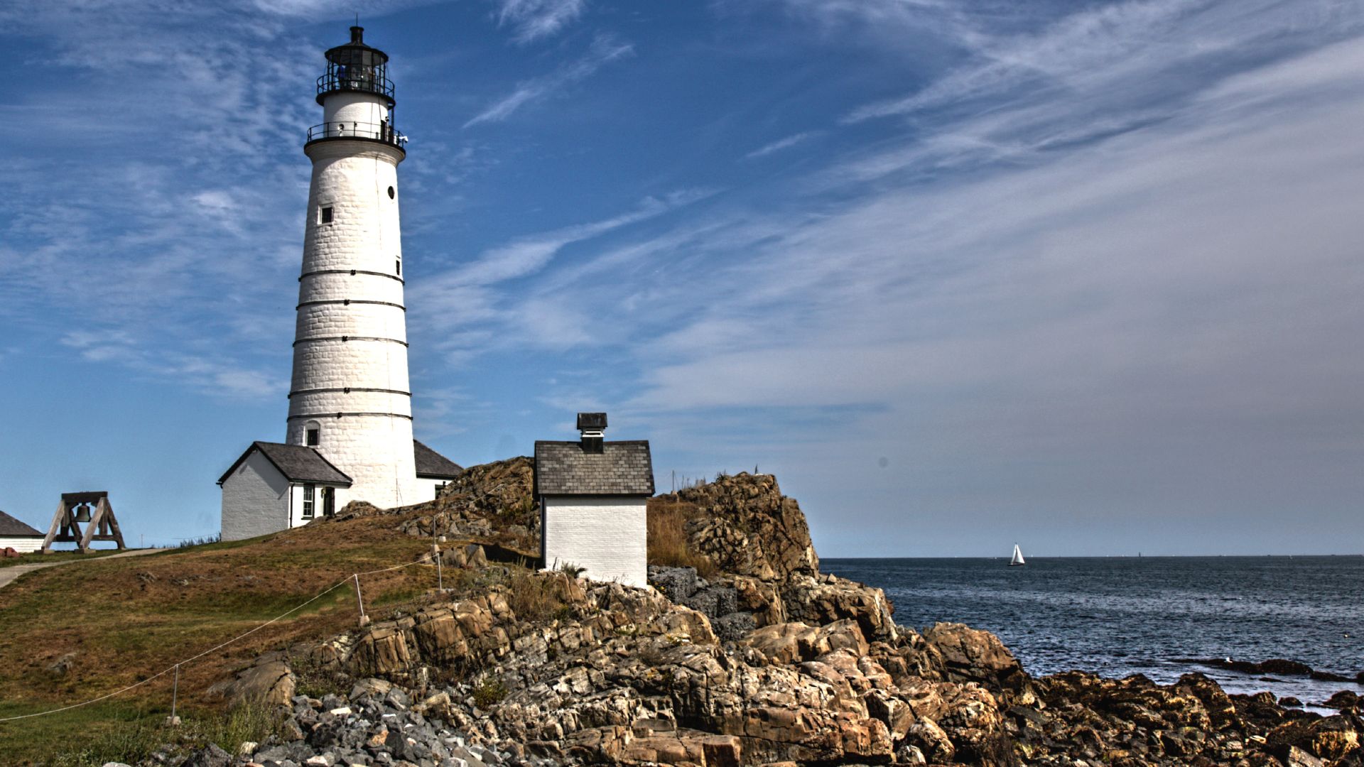 A tall, white lighthouse, known as Boston Light, stands prominently on a rocky island with sparse vegetation, overlooking the ocean under a clear blue sky with wispy clouds. A small building sits near the base of the lighthouse, and a sailboat is visible in the distance on the water.