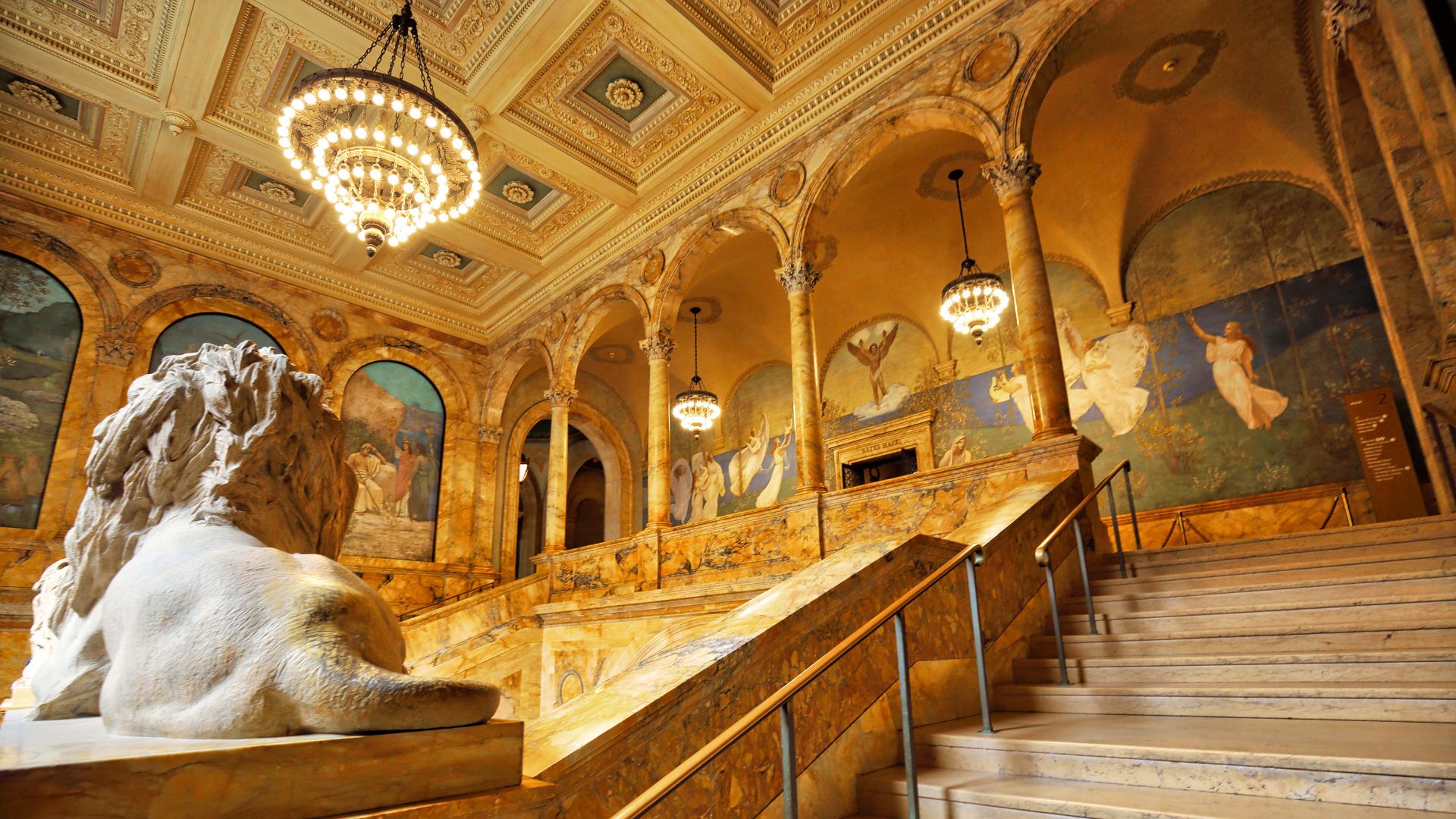 A grand, ornate interior view of the Boston Public Library's McKim Building, featuring a large marble lion sculpture in the foreground, facing a sweeping staircase leading up to arched hallways with murals, all under a high, intricately decorated ceiling with chandeliers.