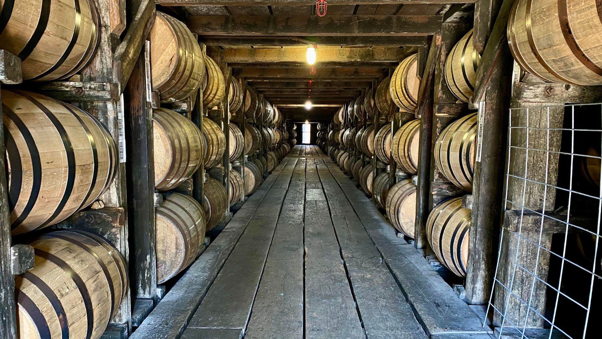 A long, narrow warehouse aisle filled with stacked wooden barrels on both sides, likely used for aging bourbon or other spirits/beers, with a wooden walkway in the center and overhead lighting.