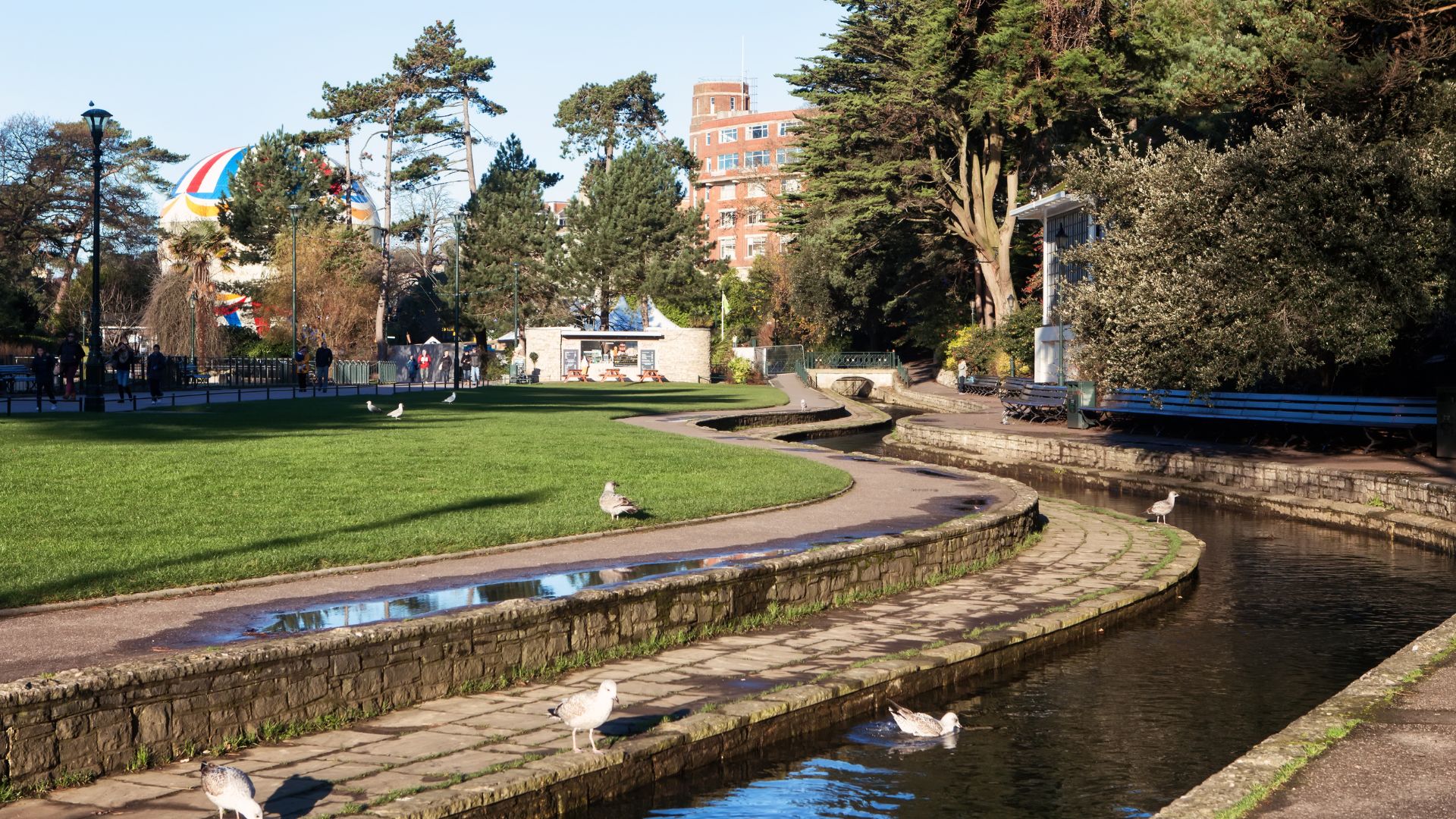 A sunny outdoor scene showing a winding stream with stone banks, a paved pathway alongside, and a grassy area with trees and benches. Several gulls are visible near the water and on the grass, and a large colorful hot air balloon is tethered in the background.
