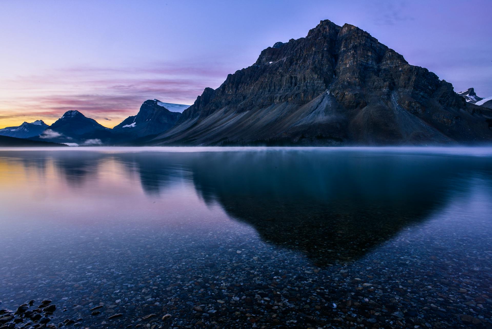 Bow Lake with its calm, turquoise waters reflecting the surrounding snow-capped mountains and evergreen forests, under a clear blue sky in the Canadian Rockies.