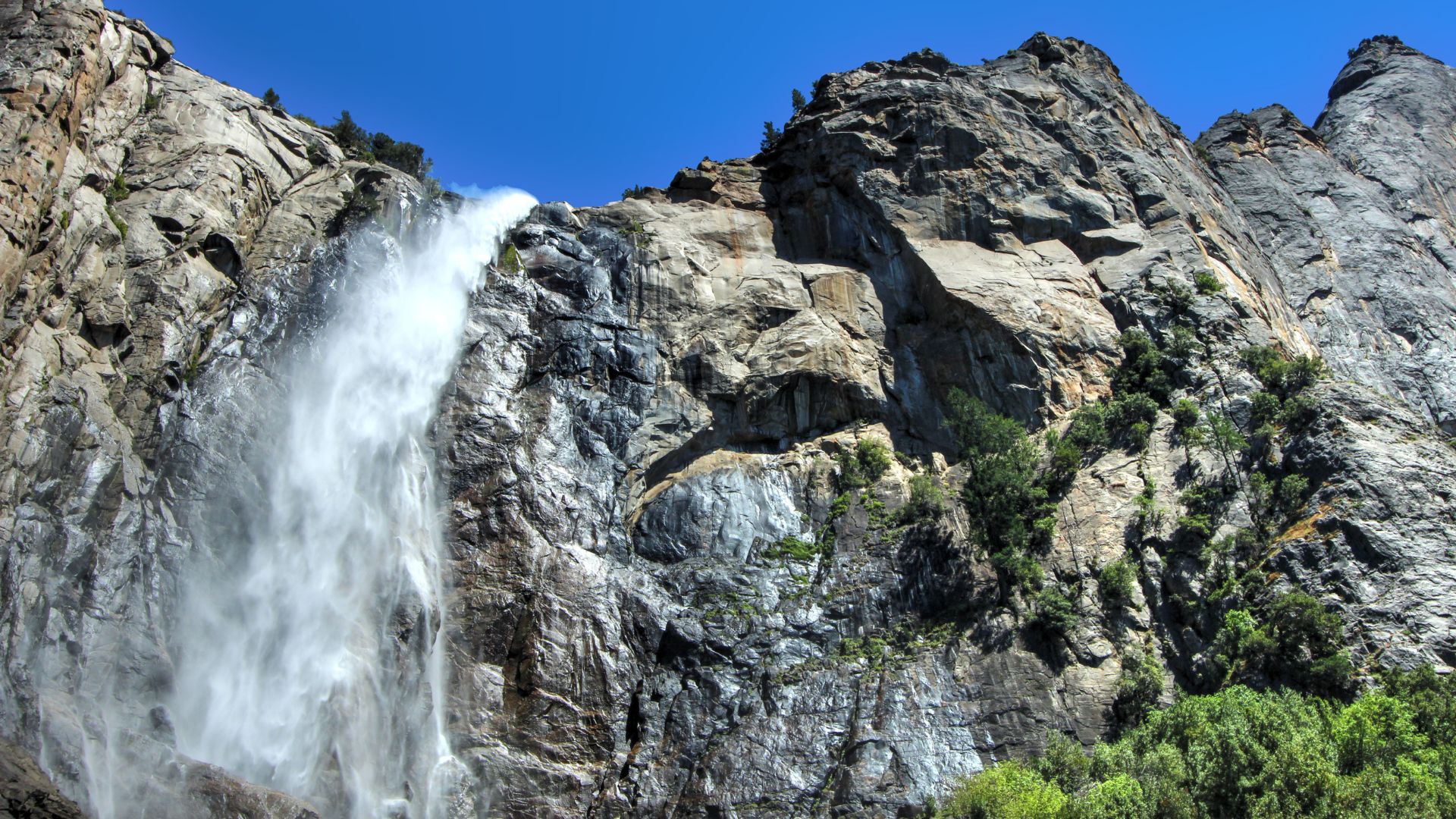 A majestic waterfall cascades down a steep, rocky cliff face under a clear blue sky, with lush green trees visible at the base of the falls and along the lower parts of the surrounding mountains.