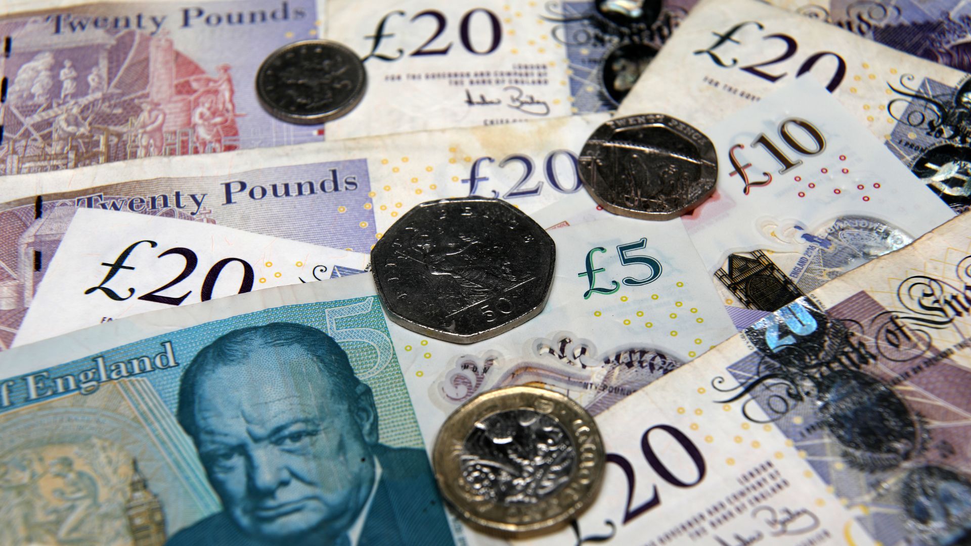A close-up shot of scattered British Pound banknotes and coins, including £20 and £5 notes, and various coins like the £2, £1, and 50p denominations.