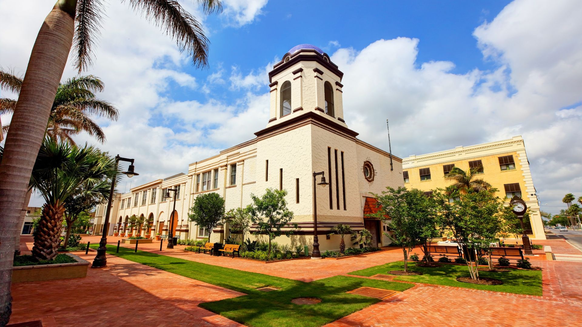 A wide-angle, sunny daytime shot of Brownsville City Hall, a large, light-colored building with a prominent central tower topped by a dark dome. The building is surrounded by green lawns and paved walkways, with palm trees and streetlights visible on the left, and a vibrant blue sky with white clouds overhead.